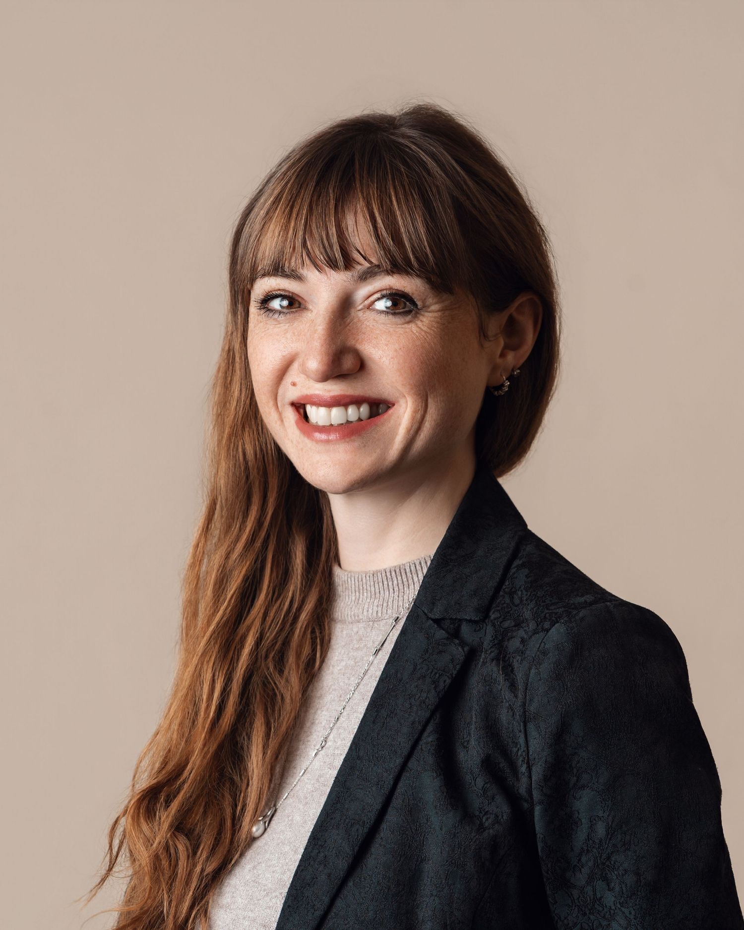 Smiling woman with bangs and long auburn hair wearing a dark blazer against a beige background.