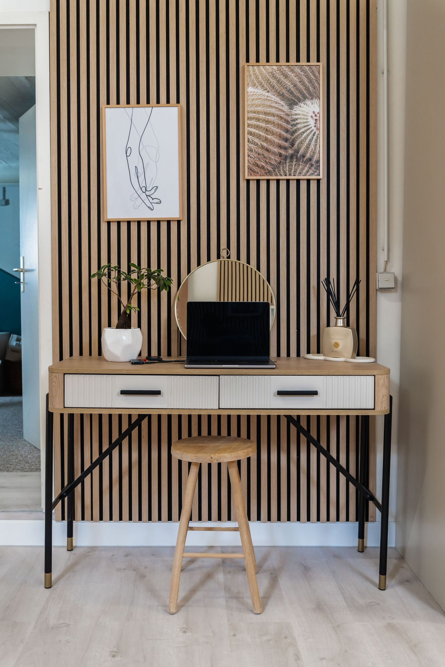 Modern Scandinavian desk with laptop, round mirror, and plant against wooden slat wall with framed art prints.