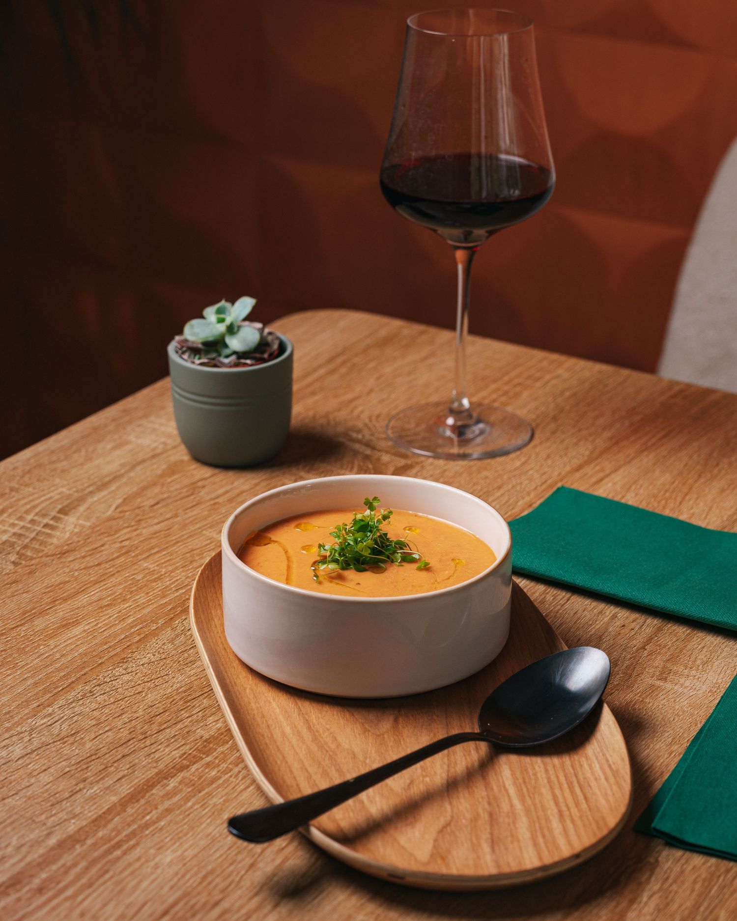 Bowl of creamy orange soup garnished with microgreens on wooden tray beside a glass of red wine.