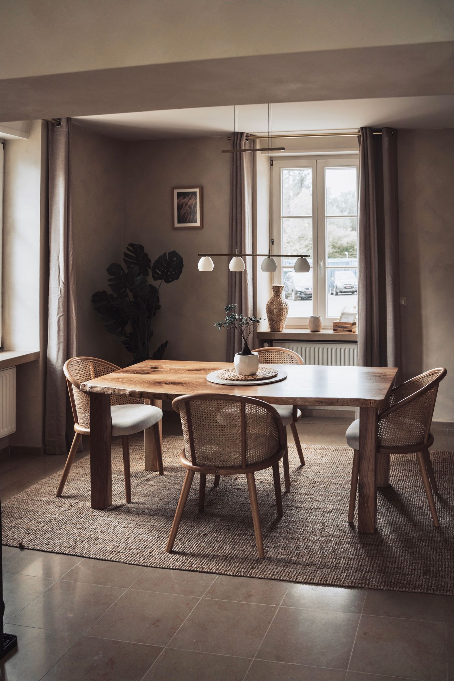 Scandinavian-style dining room with wooden table, rattan chairs, jute rug, and monstera plant near window.