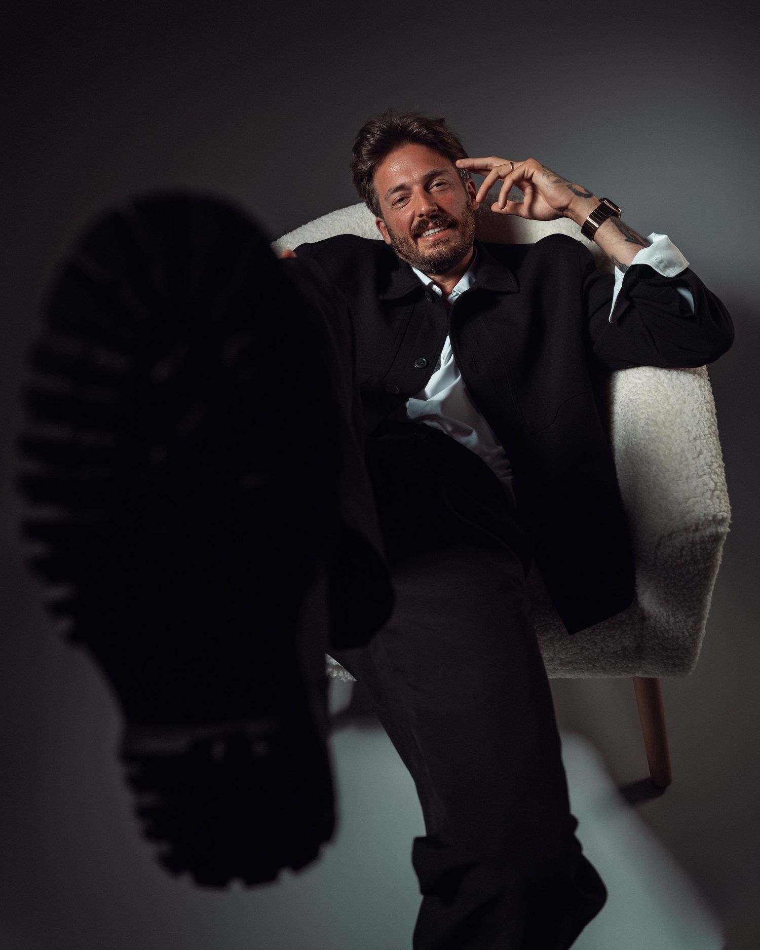 Man in black suit sitting in white chair with boot sole facing camera in dramatic studio portrait.