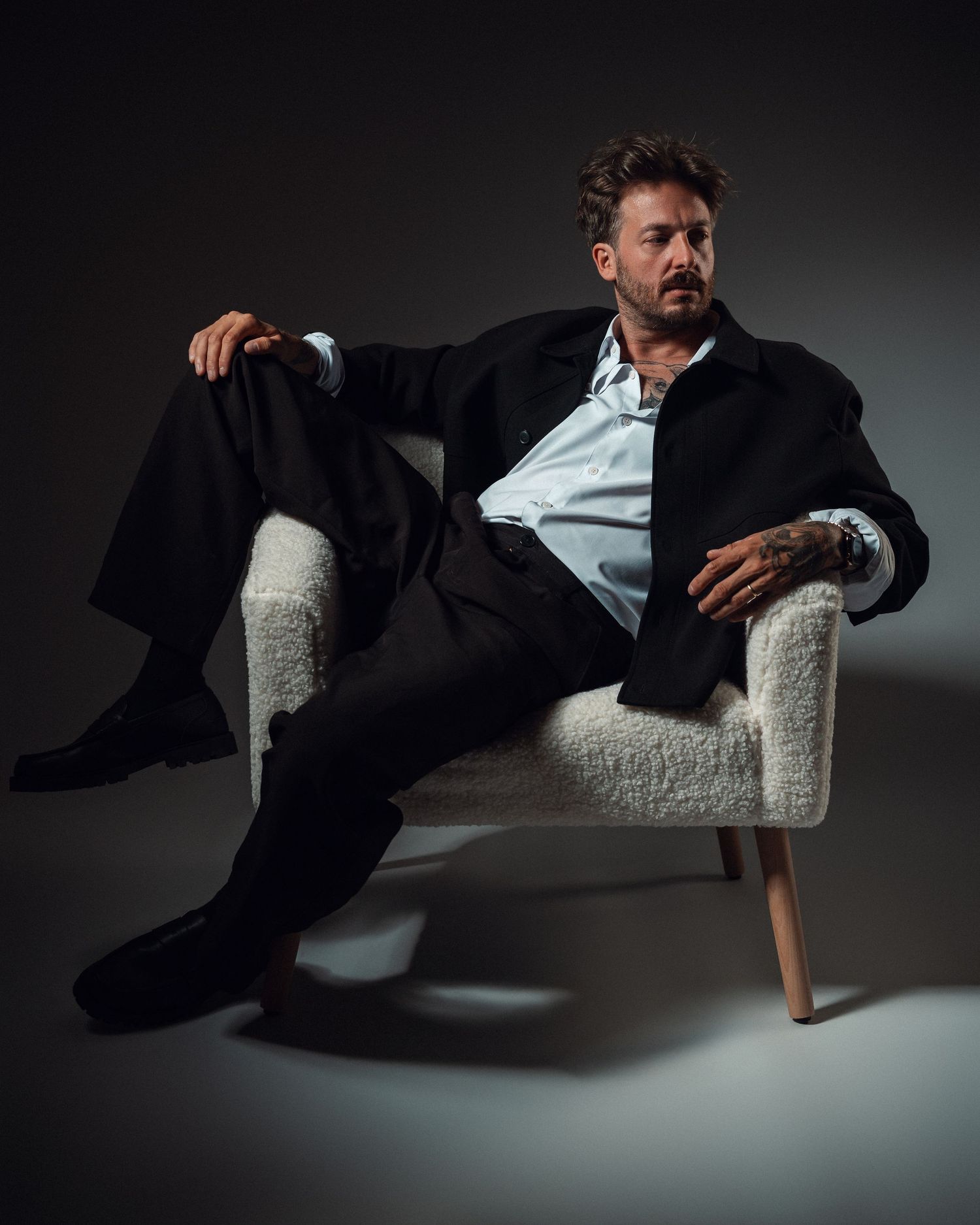 A stylish man in a black suit sits relaxed in a white boucle chair against a dark studio background.