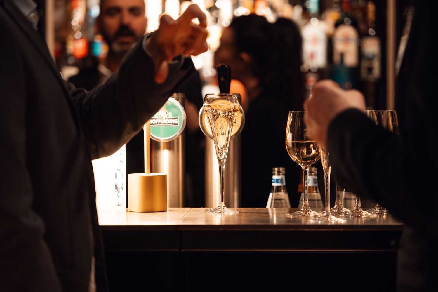Bartender pouring draft beer at a dimly lit bar with wine glasses and bottles on the counter.