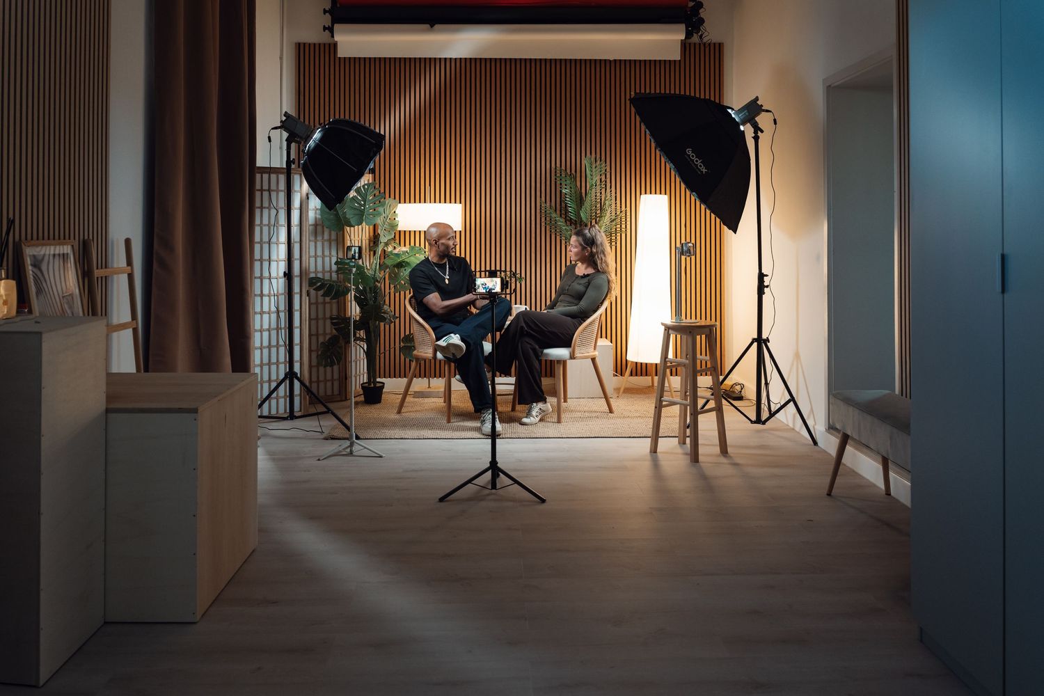 Two people sit in a professional photo studio setup with softbox lights and camera equipment on tripods.