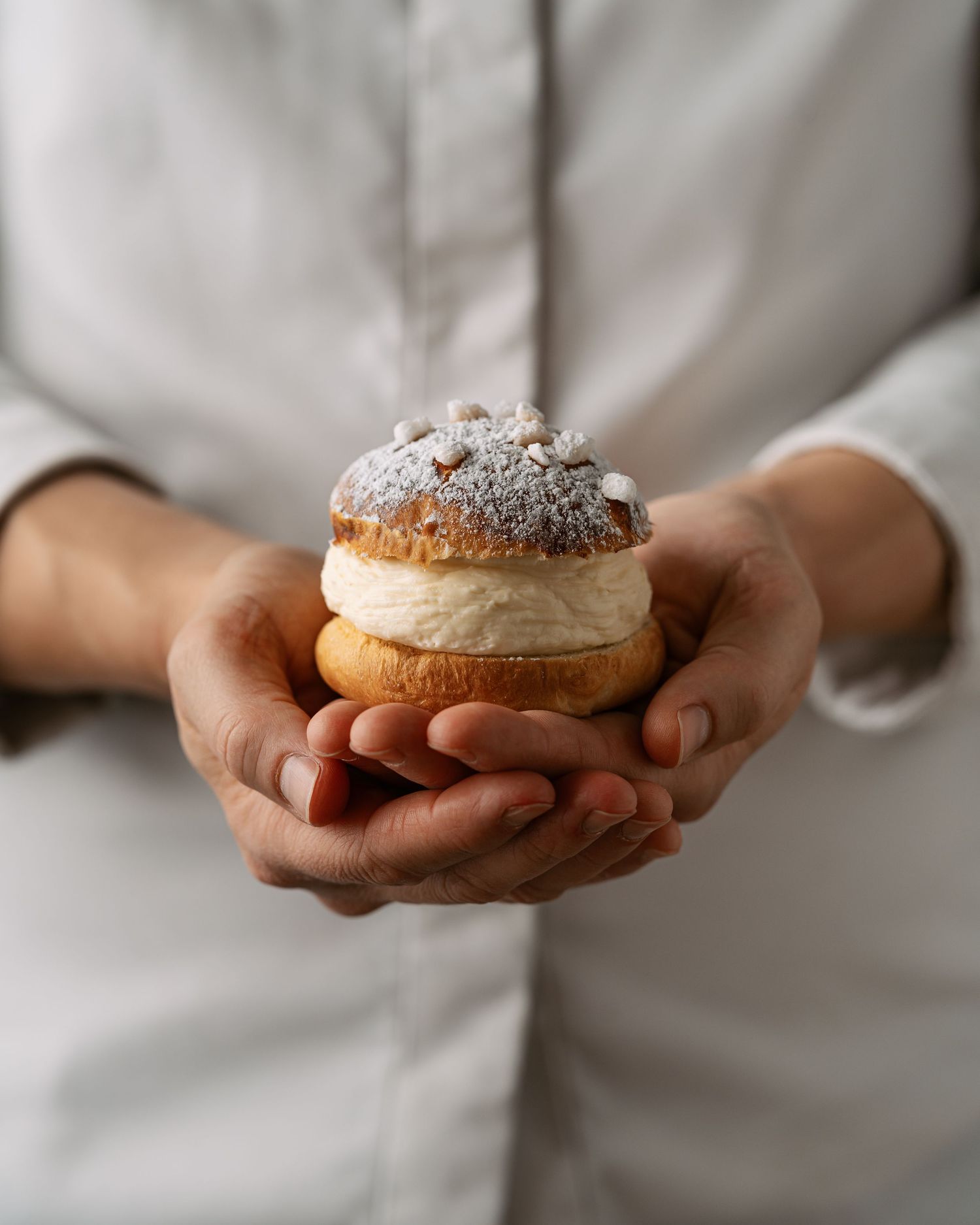 Baker in white uniform holding a powdered sugar-topped cream-filled brioche bun with both hands.