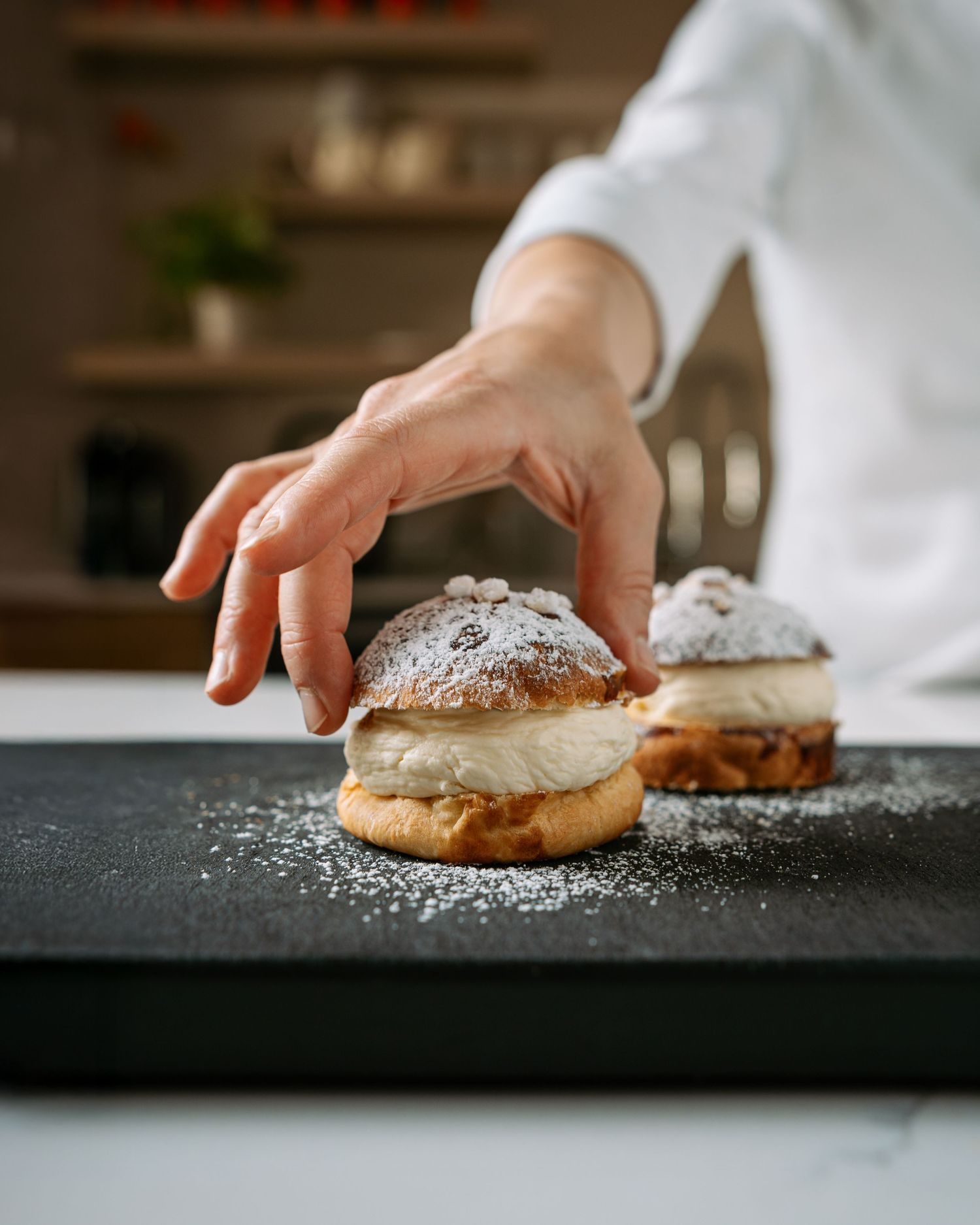 Chef placing powdered sugar-dusted cream-filled buns on a dark slate board in a kitchen.
