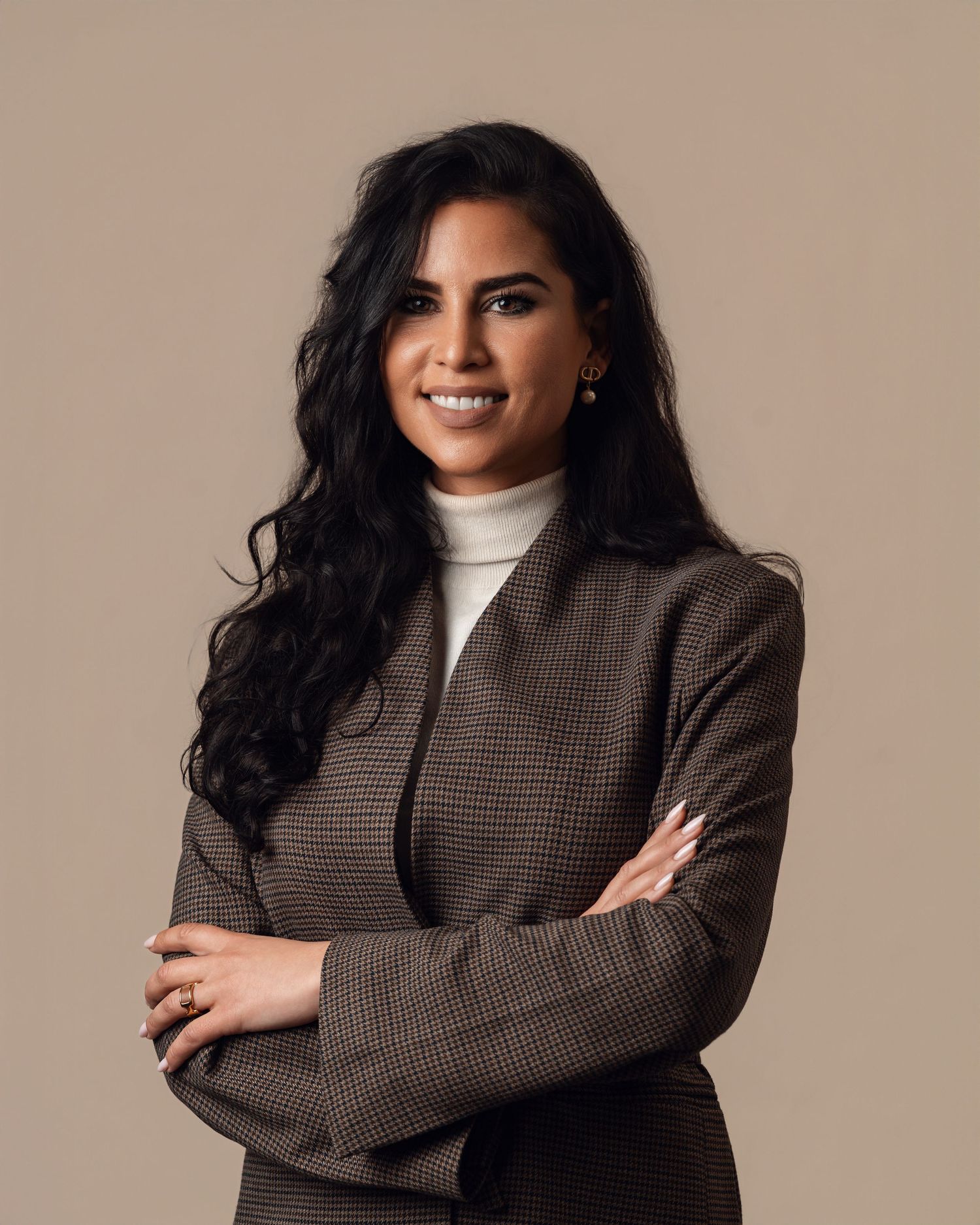 Professional woman in a plaid blazer with arms crossed smiling against a neutral beige background.