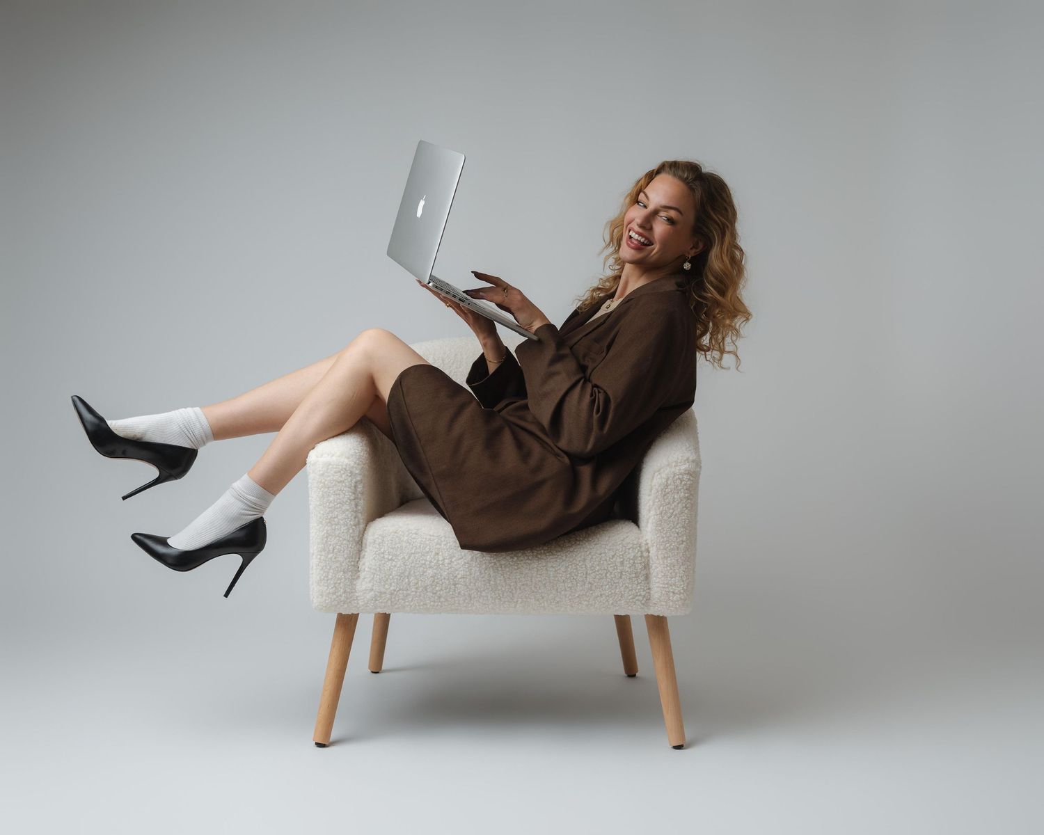 Woman in brown dress sitting in white chair with laptop, wearing black heels and white socks, smiling at camera.
