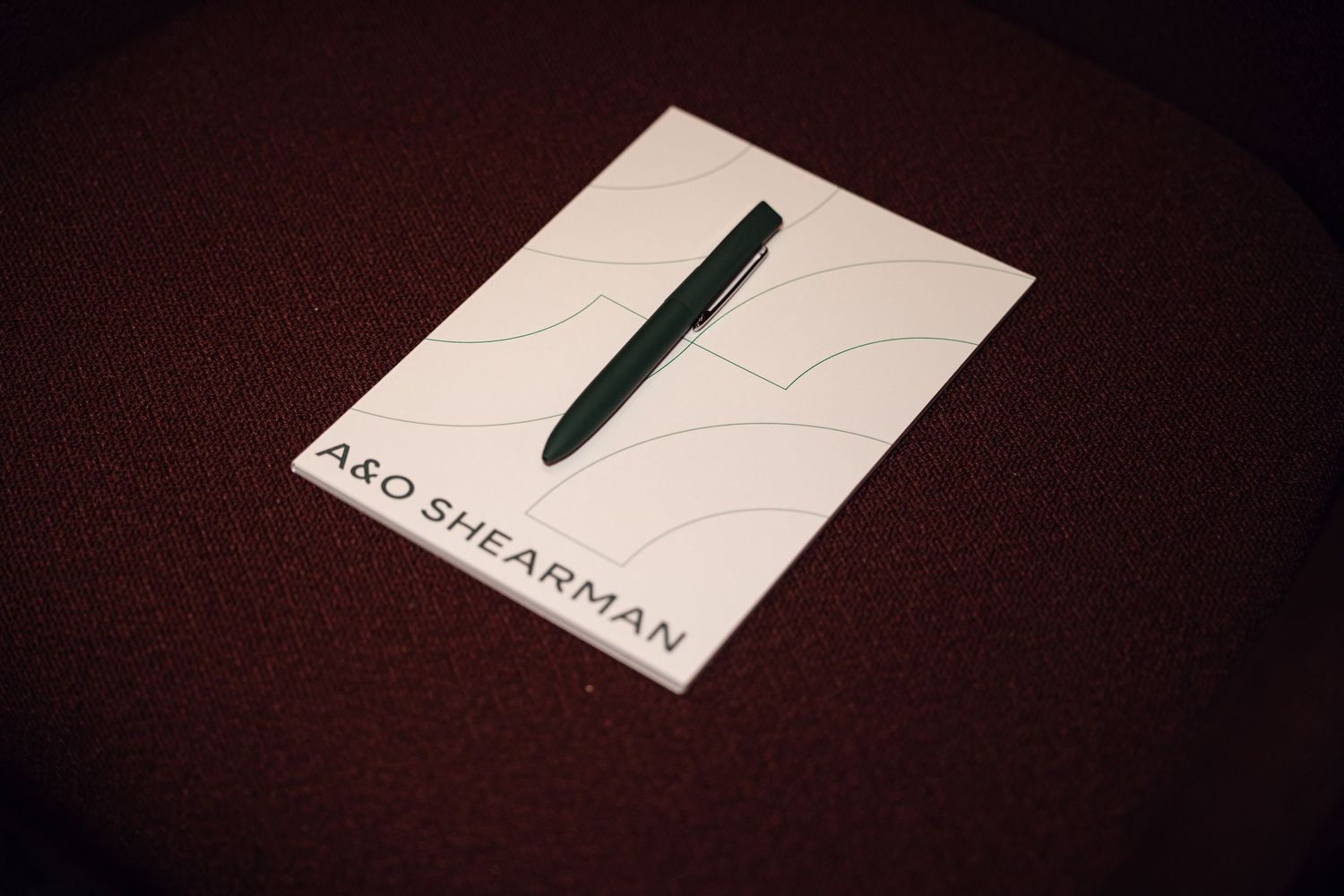 A&O Shearman branded notepad with green pen on dark red textured surface.