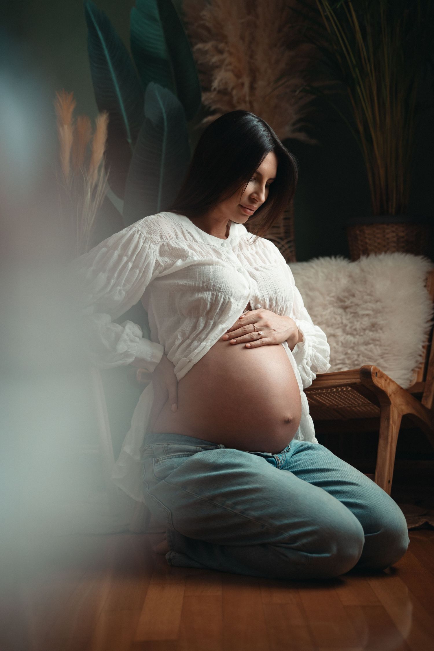 Pregnant woman in white blouse cradles her bare belly while kneeling on wooden floor.