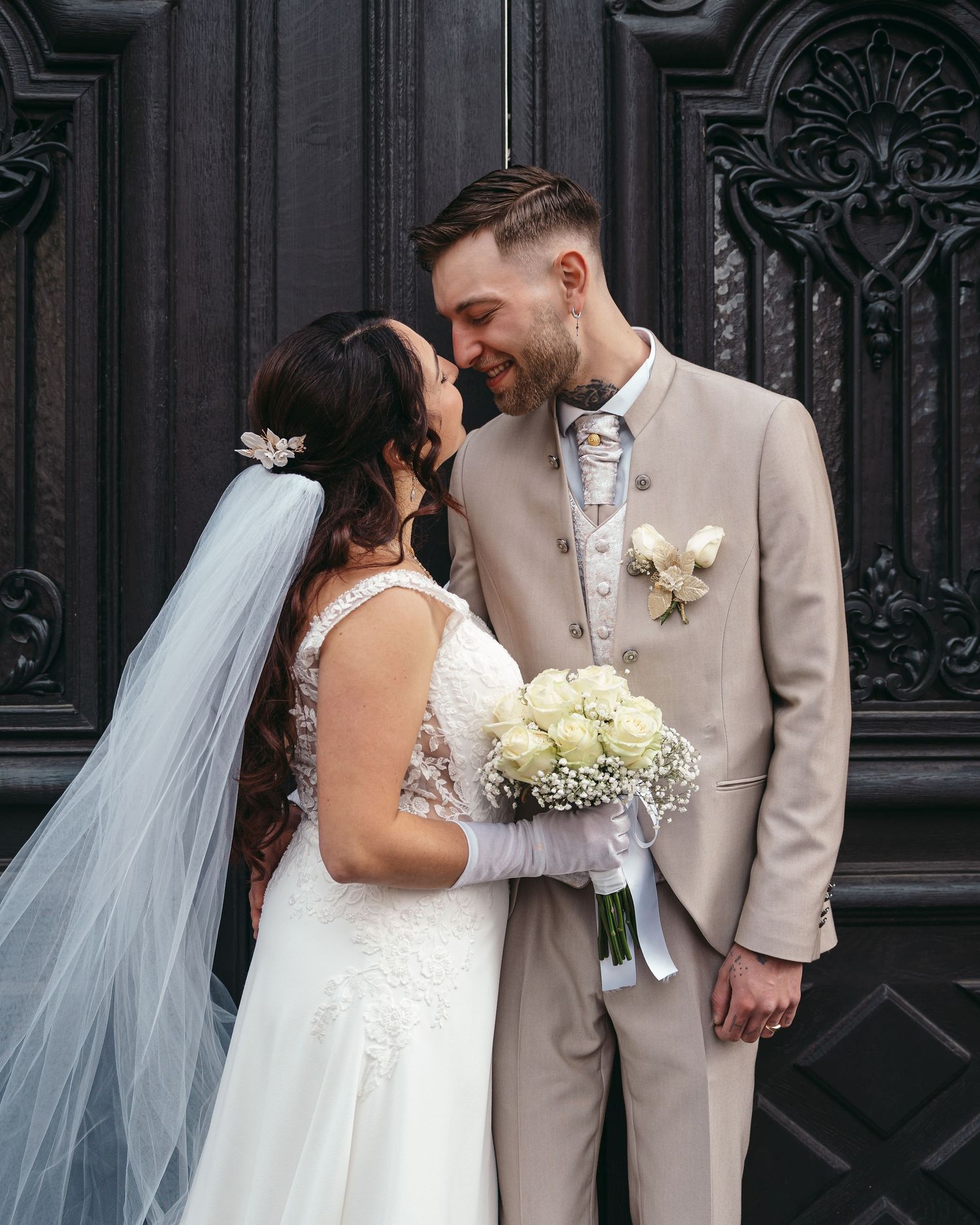 Bride and groom share a romantic moment in front of ornate dark doors, bride holding white rose bouquet wearing veil.