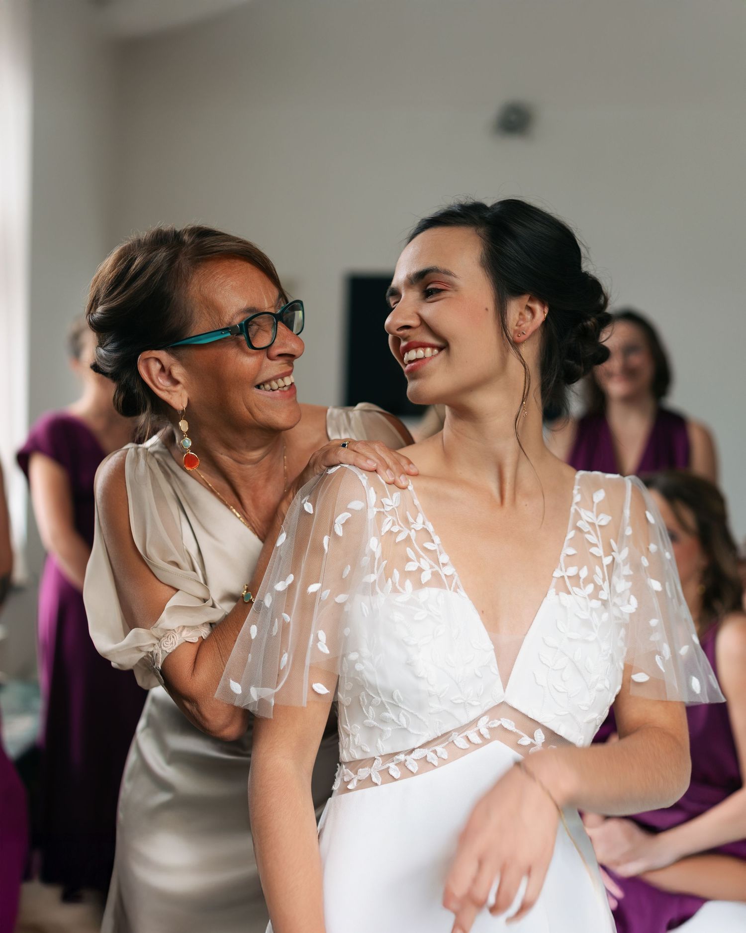Bride in white lace gown laughs joyfully with older woman in glasses during wedding preparations.