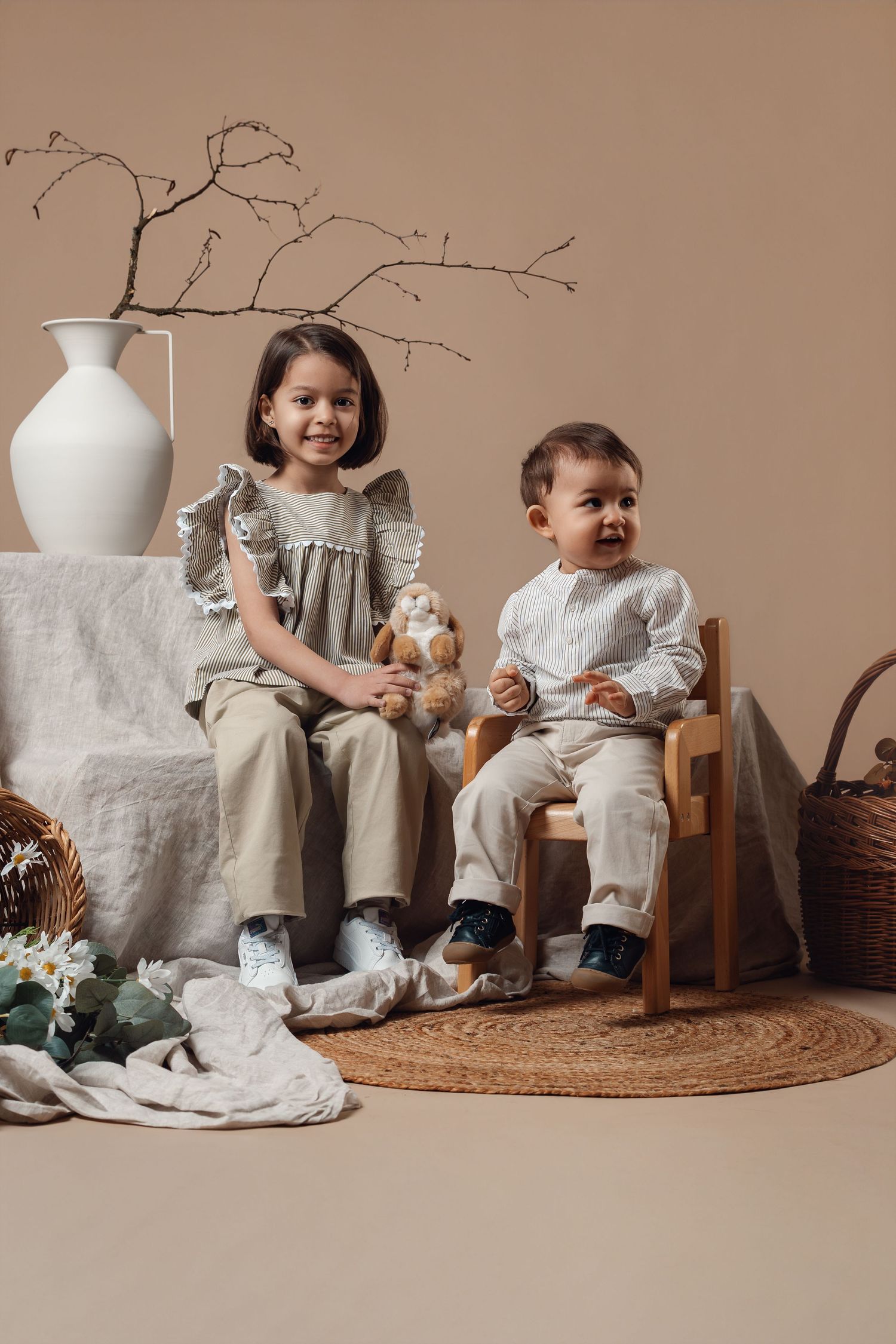 Two young children in neutral linen outfits sit together in a styled studio setting with a white vase and wicker basket.