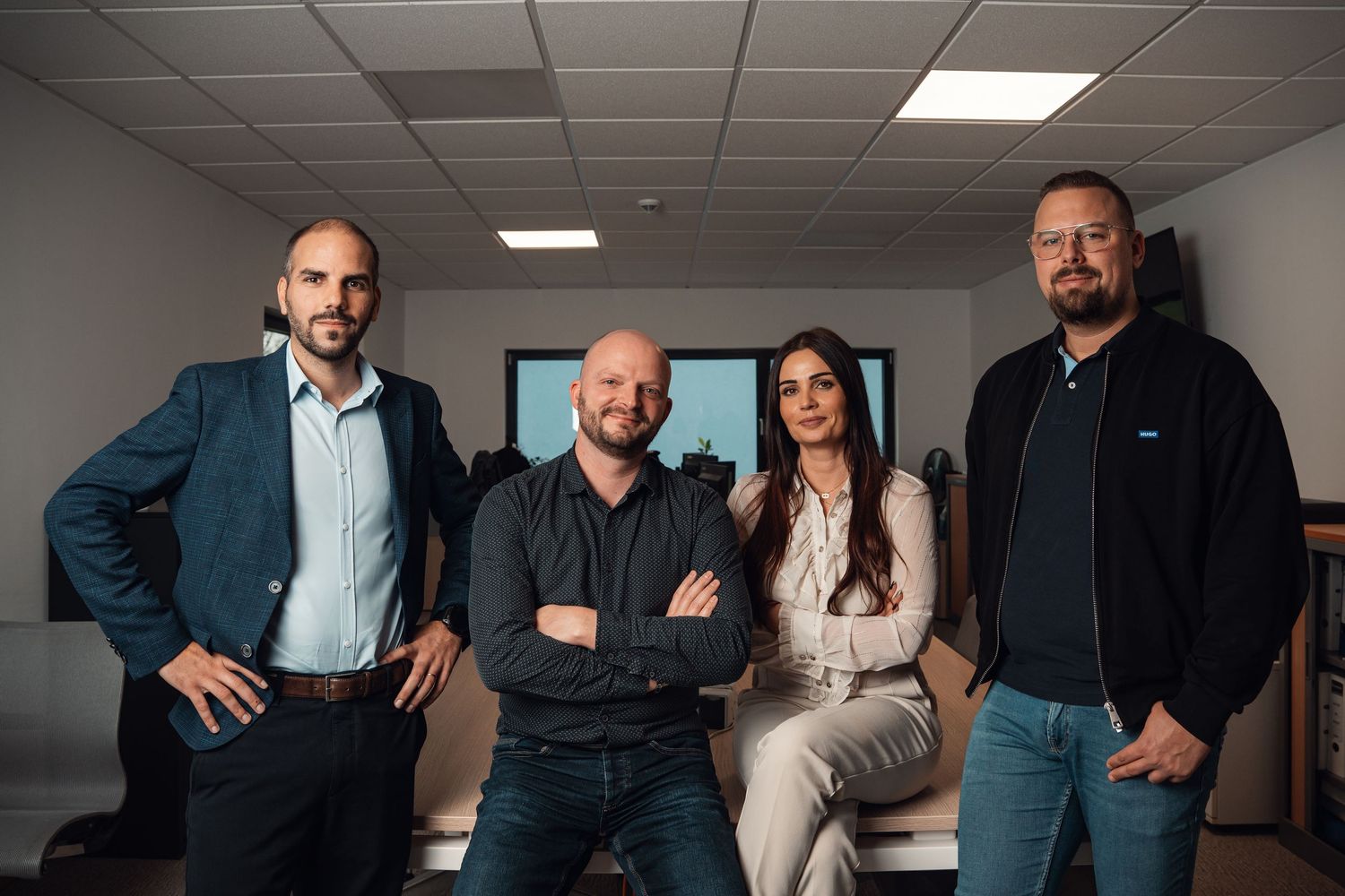 Four business professionals posing together in a modern office setting, two men and one woman standing with one seated.