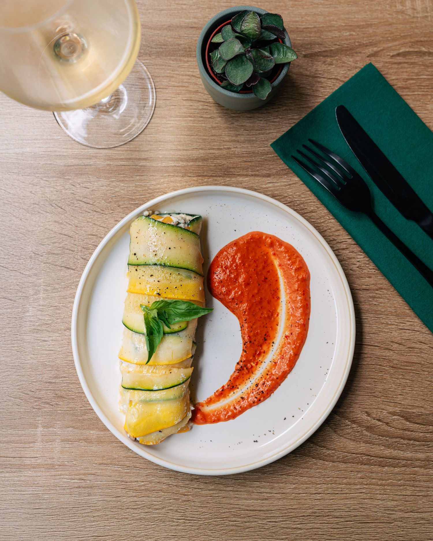Zucchini roll with red pepper sauce on a white plate beside white wine and green napkin.