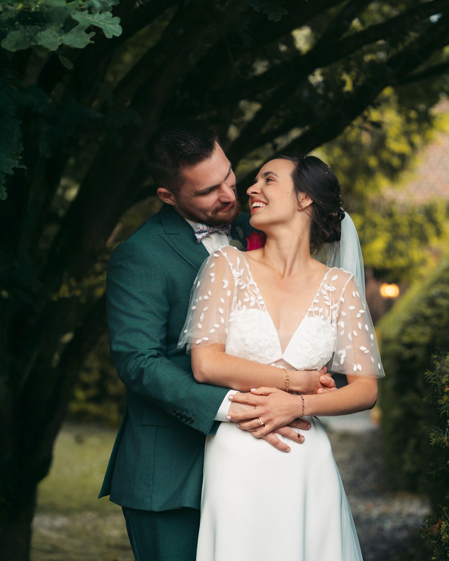 Bride and groom embrace tenderly under lush green trees in golden evening light.