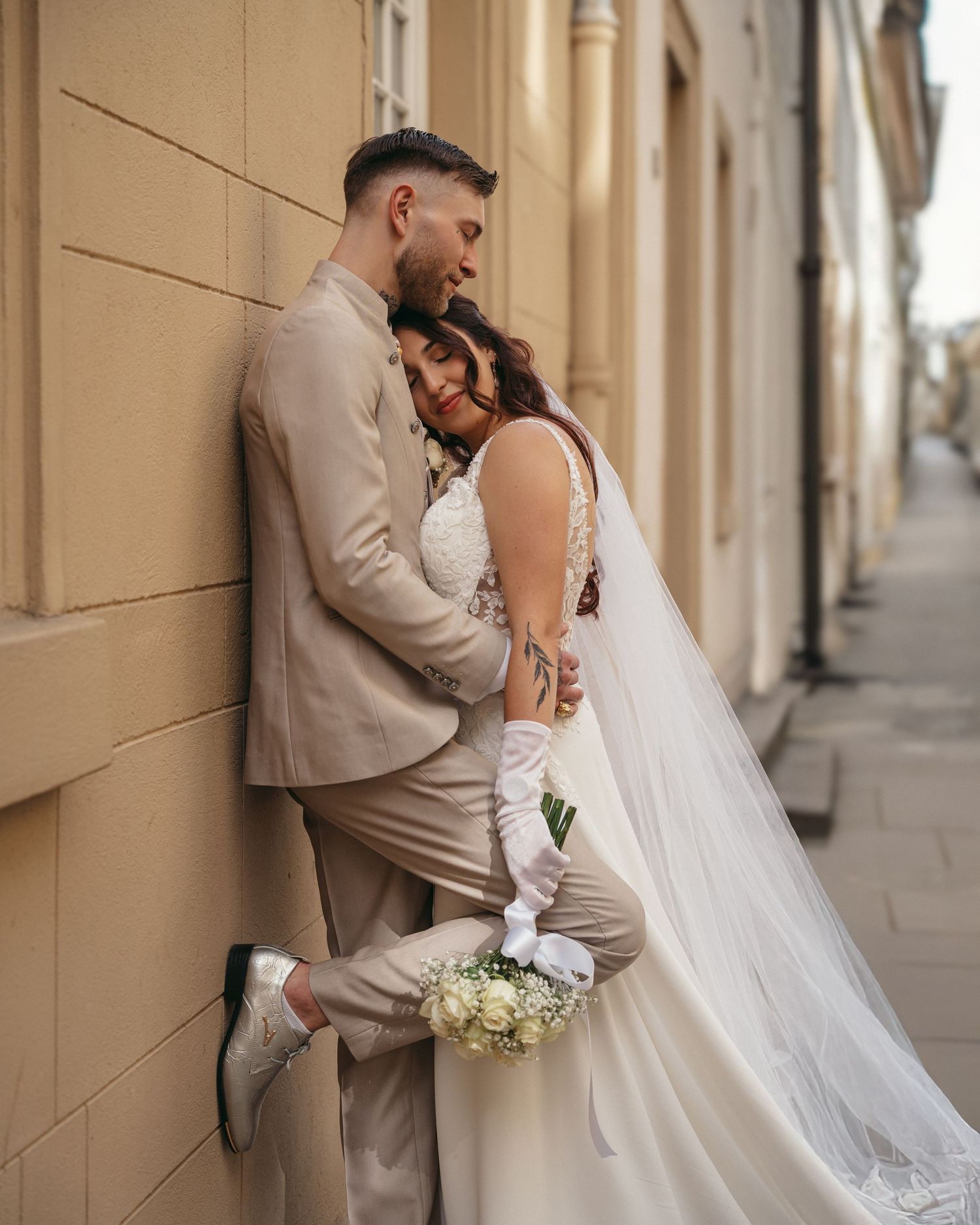 Bride and groom embrace against a beige building wall, bride in white gown with veil holding bouquet, groom in tan suit.