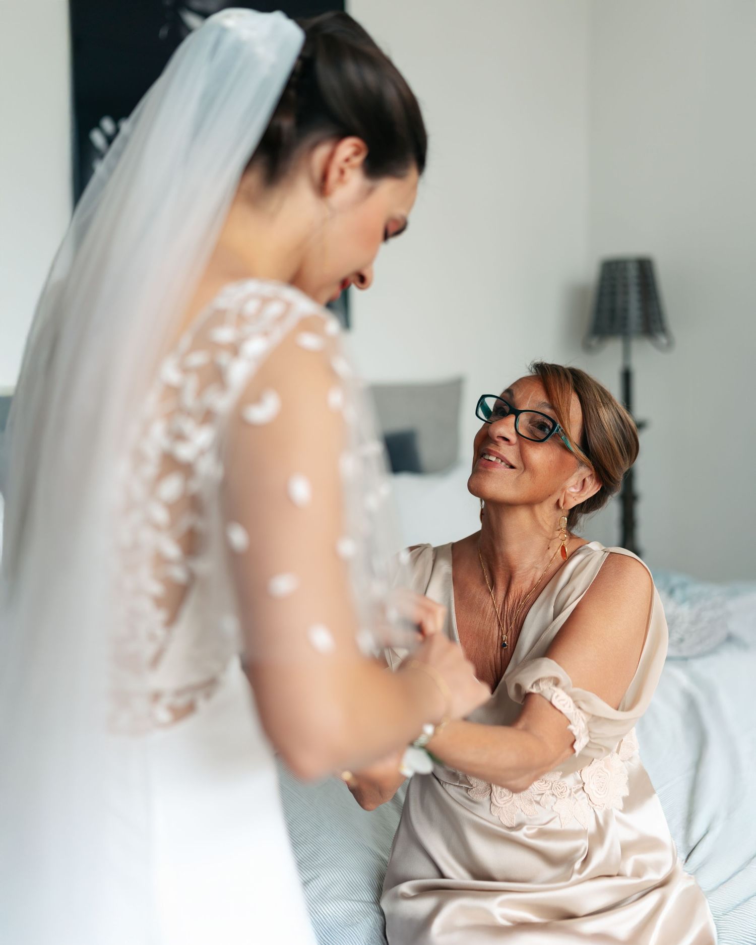 Mother kneels smiling while helping bride in white lace veil with wedding dress preparations.