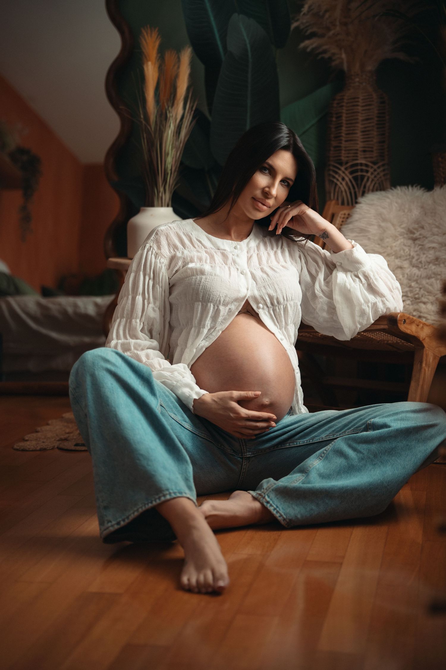 Pregnant woman in white blouse and jeans sitting on wooden floor, cradling baby bump in cozy boho-style room.