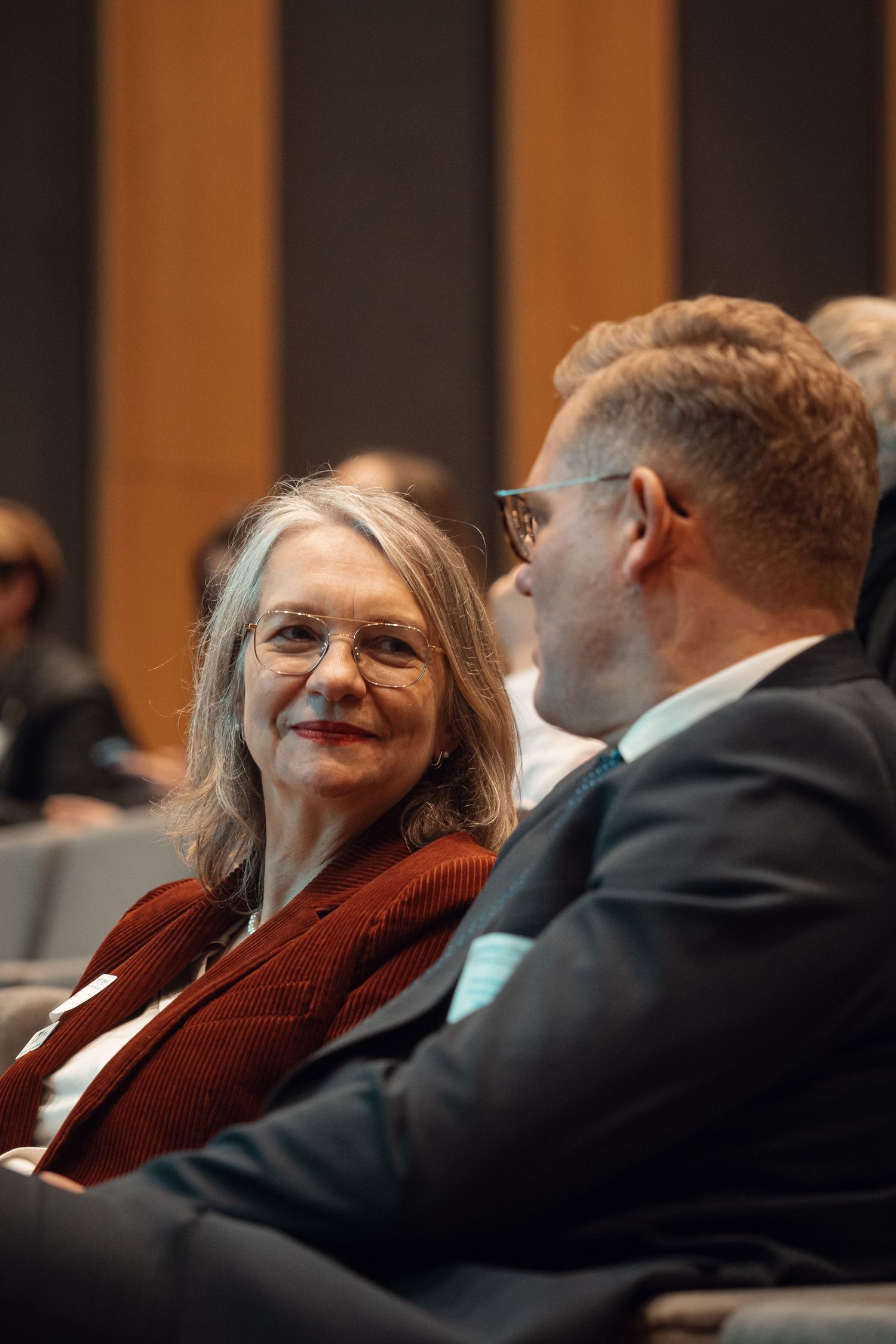 A woman with gray hair and glasses smiles at a man in a suit during a professional conference.