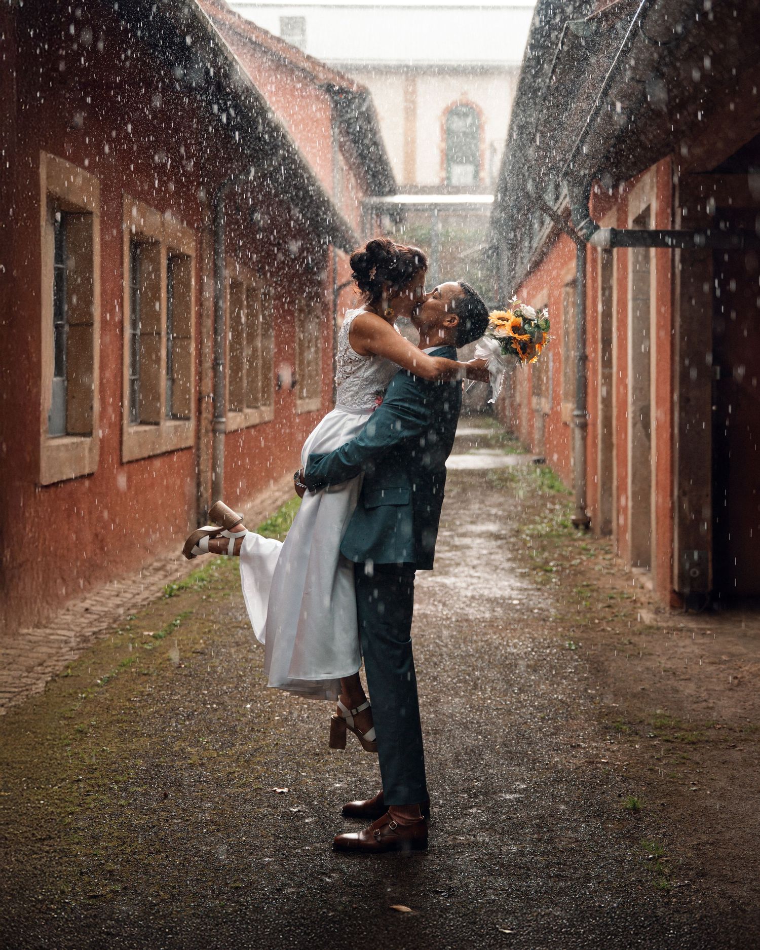 Bride and groom share a romantic kiss in the rain, with groom lifting bride in a cobblestone alley.