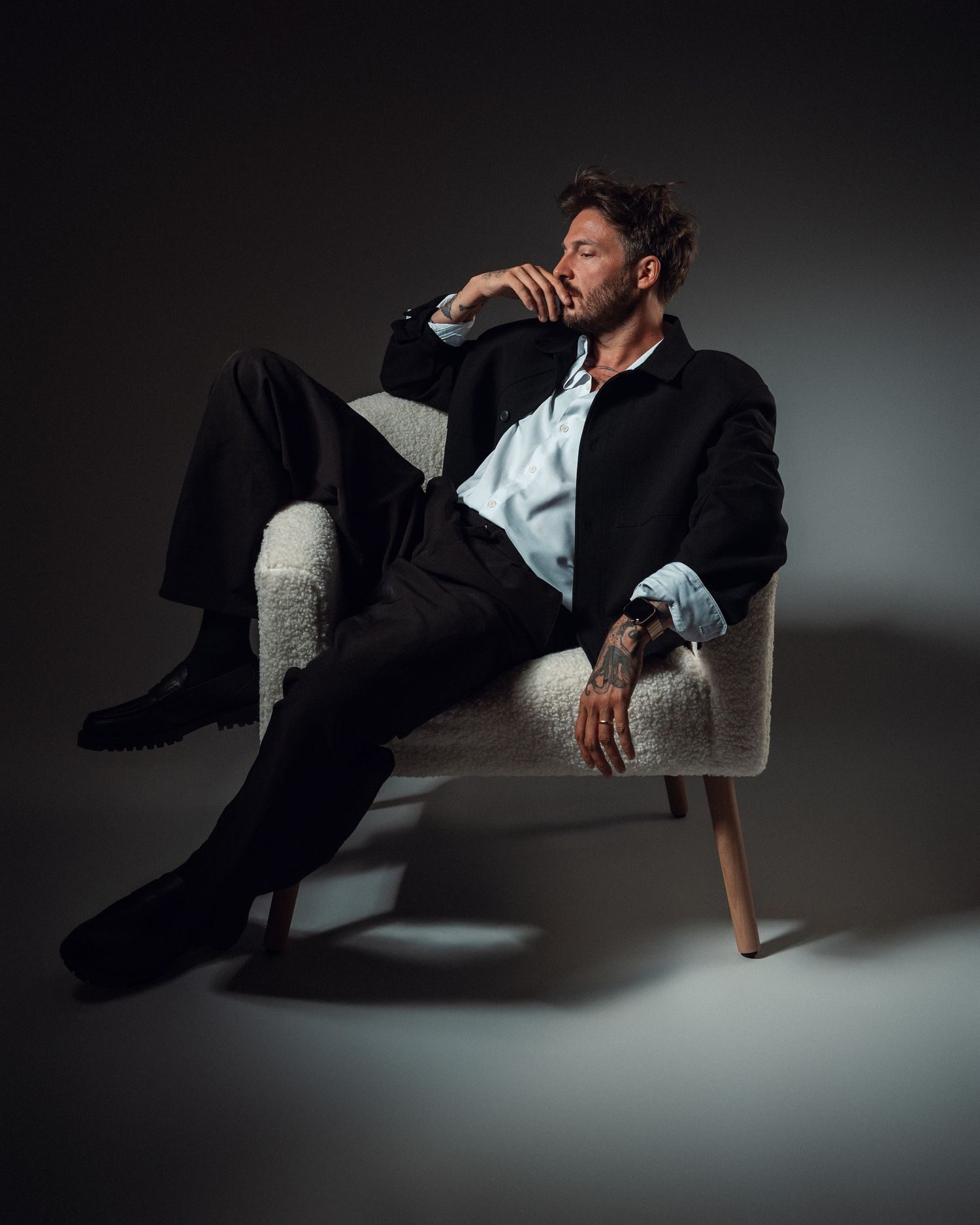 Man in black jacket and trousers sitting relaxed in a white textured armchair, studio portrait with dramatic lighting.