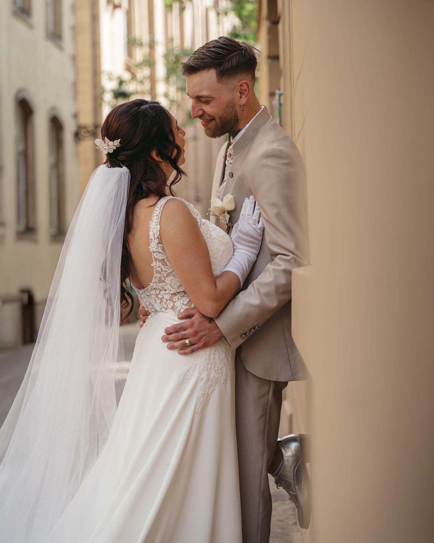 A bride and groom share a romantic moment in a sunlit European alley, her white gown and veil flowing elegantly.