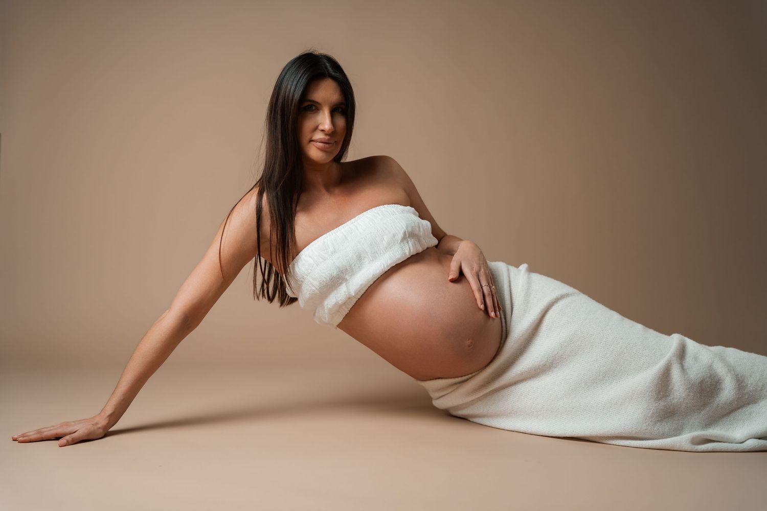 Pregnant woman posing gracefully wrapped in white fabric on beige background.