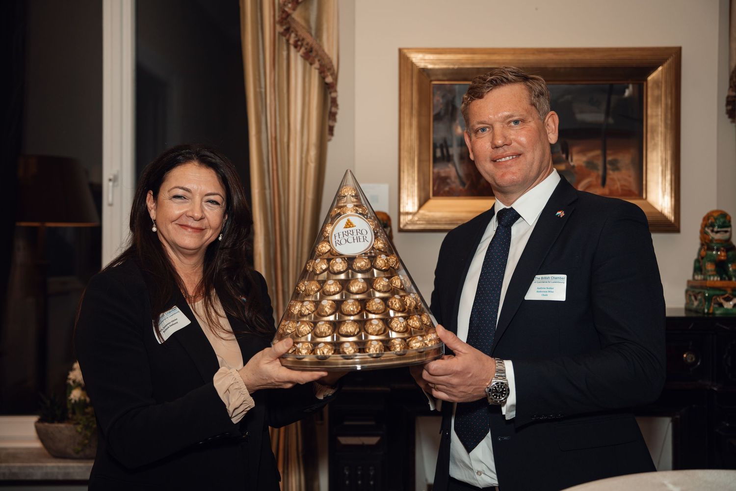 Two professionals smiling while holding a large Ferrero Rocher chocolate pyramid display at a formal business event.