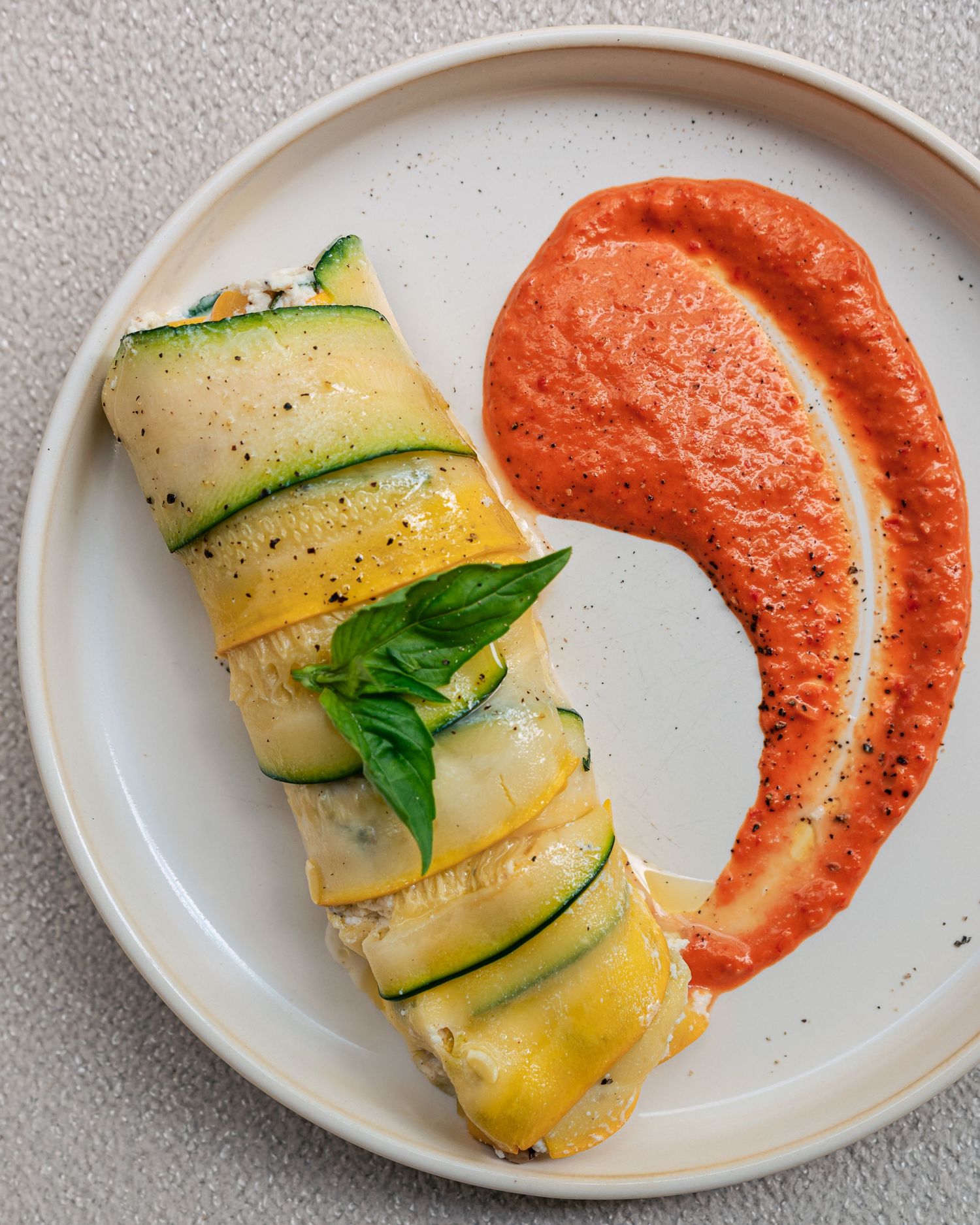 Elegant zucchini roll stuffed with cheese served with roasted red pepper sauce on a white plate.