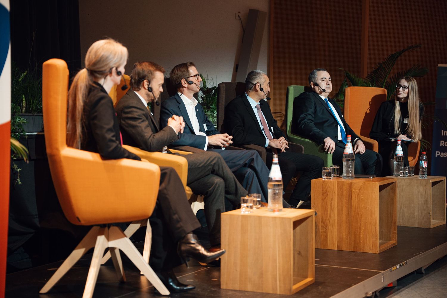 Panel of business speakers engaged in discussion seated in orange chairs at corporate conference.