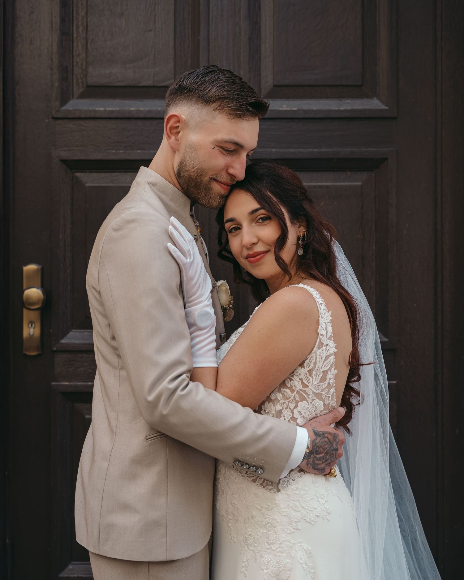 Couple embracing in wedding attire before dark wooden door, groom in beige suit, bride in lace gown with veil.