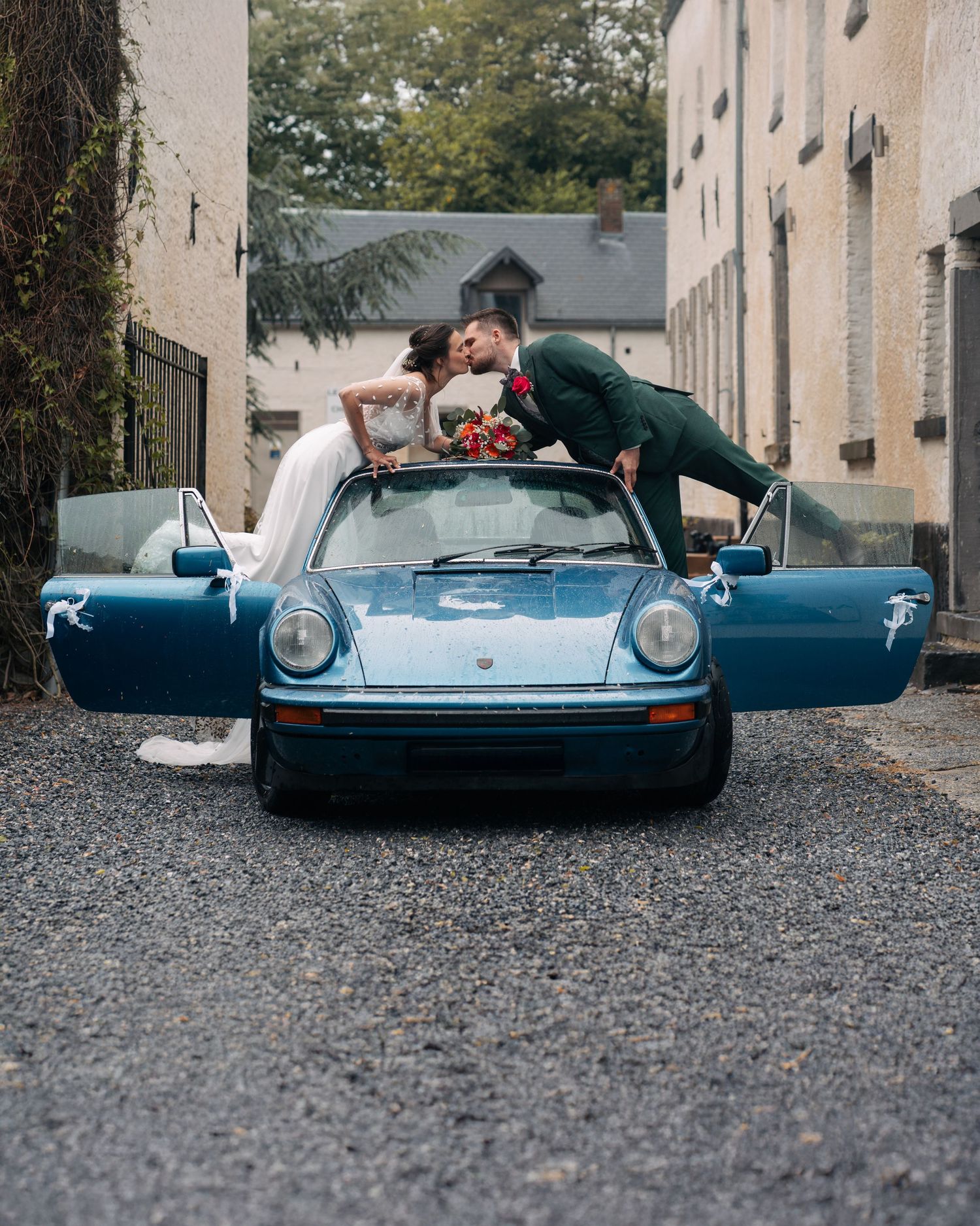 Newlyweds kissing over a classic blue Porsche 911 convertible with doors open in a courtyard.