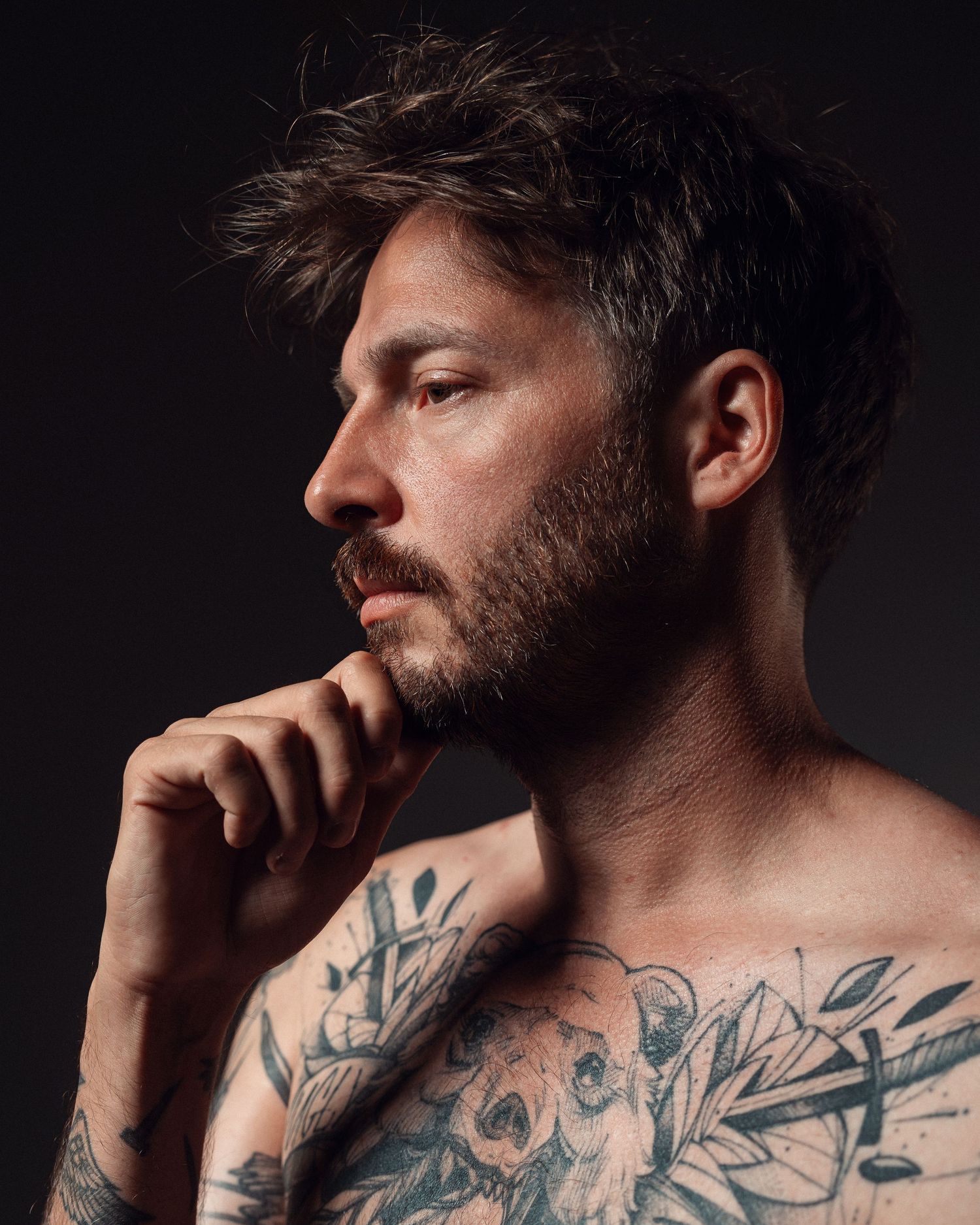 Shirtless tattooed man with beard in thoughtful pose against dark background, hand on chin.