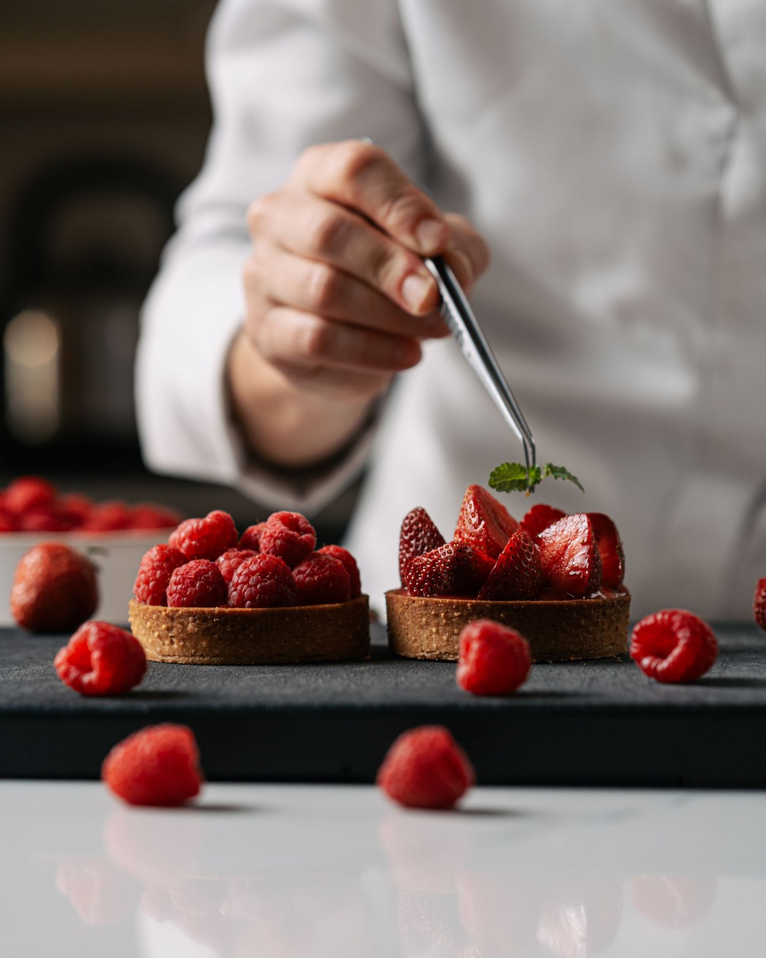 Chef garnishing fresh raspberry and strawberry tarts on a dark slate board with scattered berries.