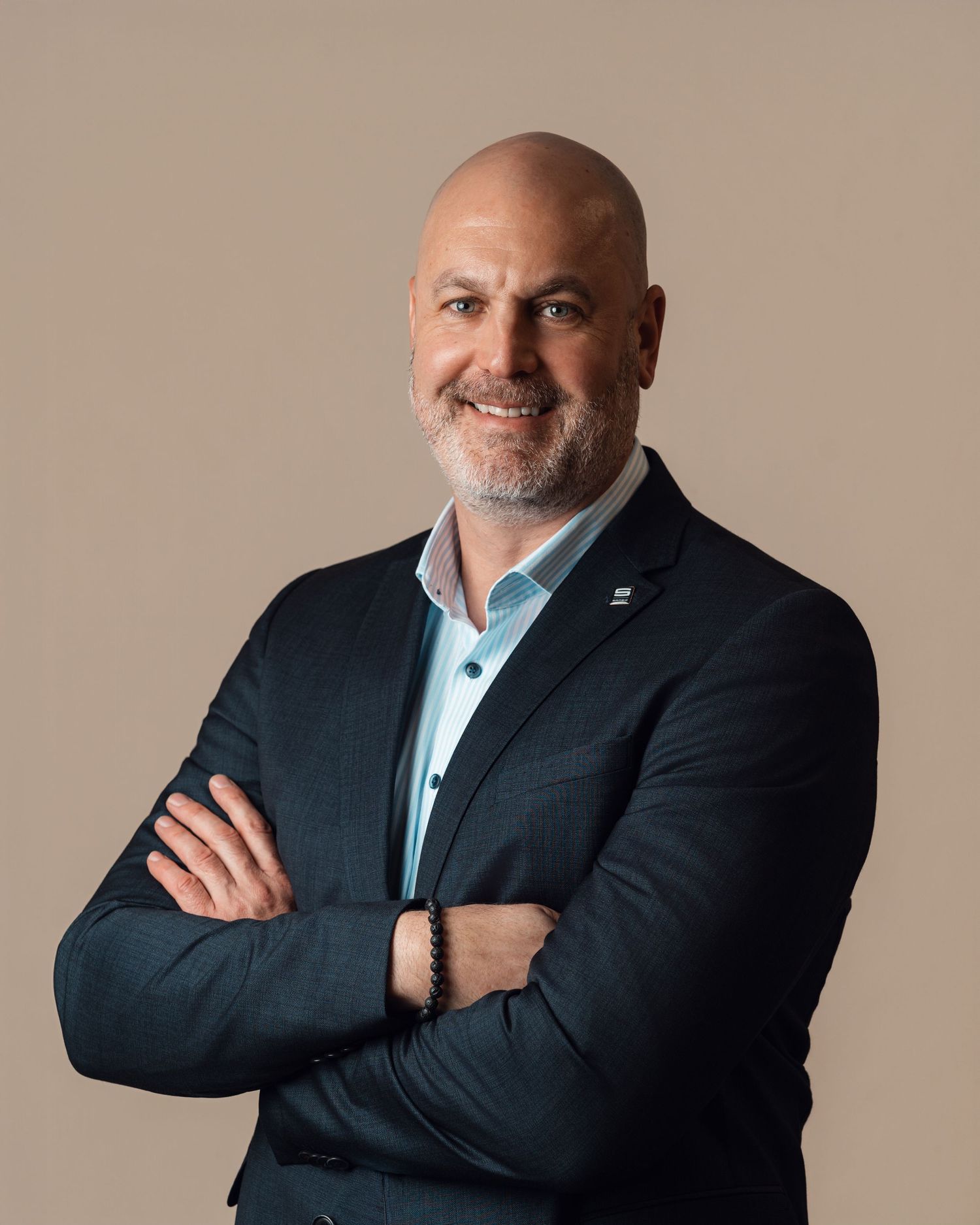 Bald professional man with a beard smiling, wearing a dark blazer with arms crossed against a warm beige background.