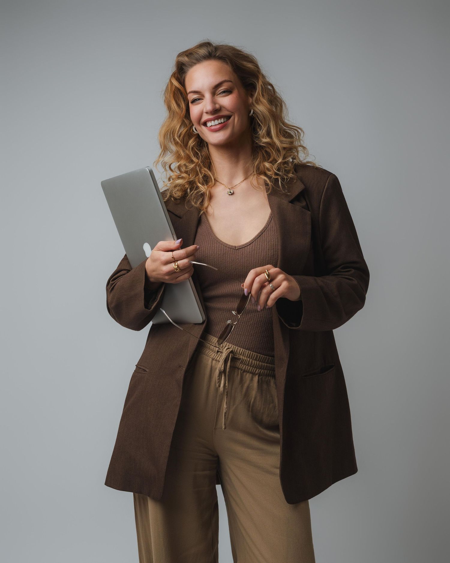 Smiling woman in brown blazer and tan pants holding a laptop and glasses against a gray background.