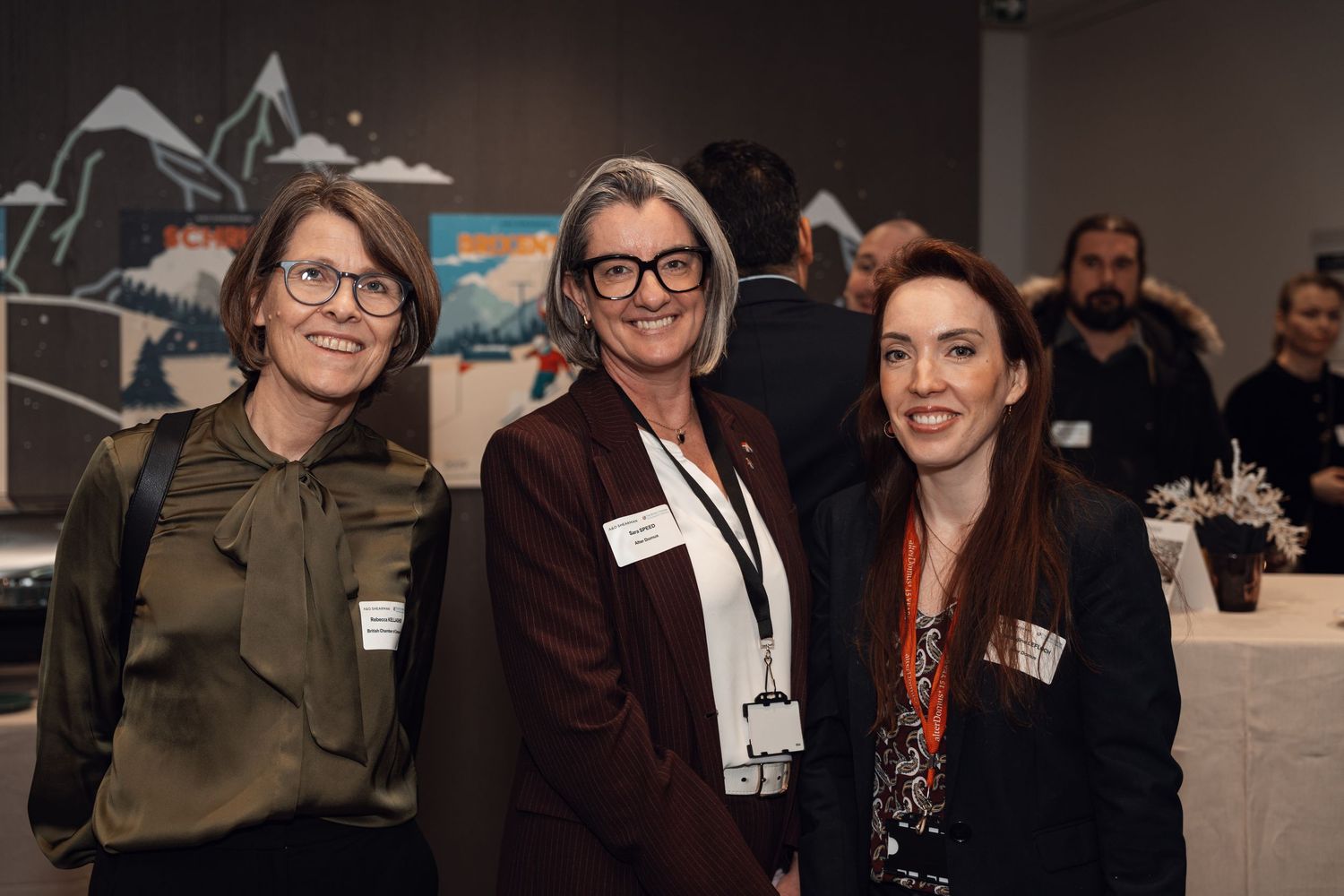 Three professional women smiling together at a corporate networking event with name badges.