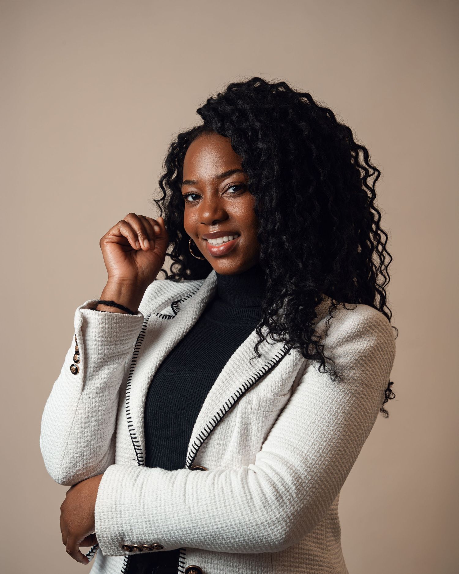 Confident Black woman with curly hair smiling, wearing a white blazer over a black turtleneck against a beige background.