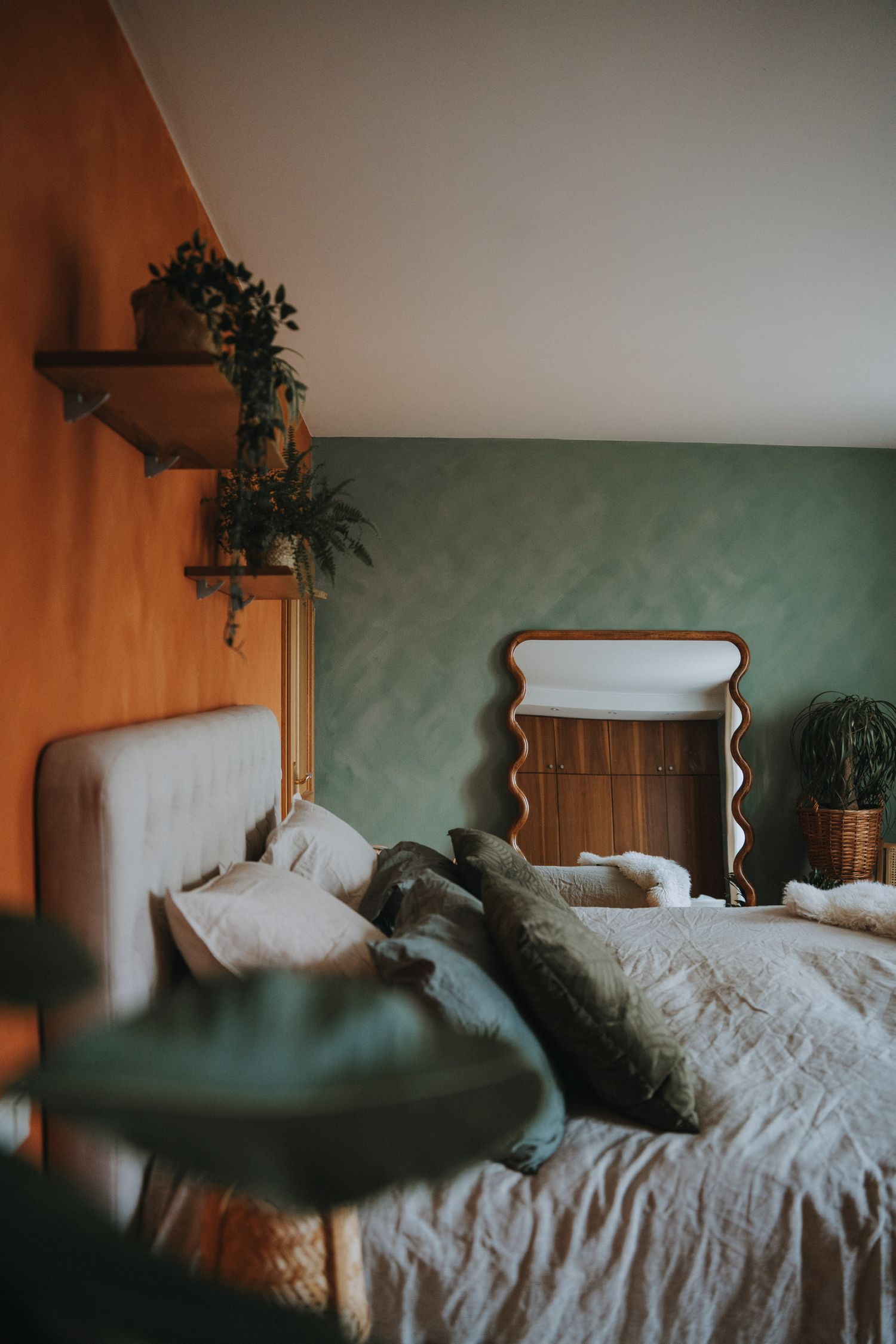 Cozy bedroom with orange and green walls, white upholstered bed, decorative mirror, and potted plants on wooden shelves.