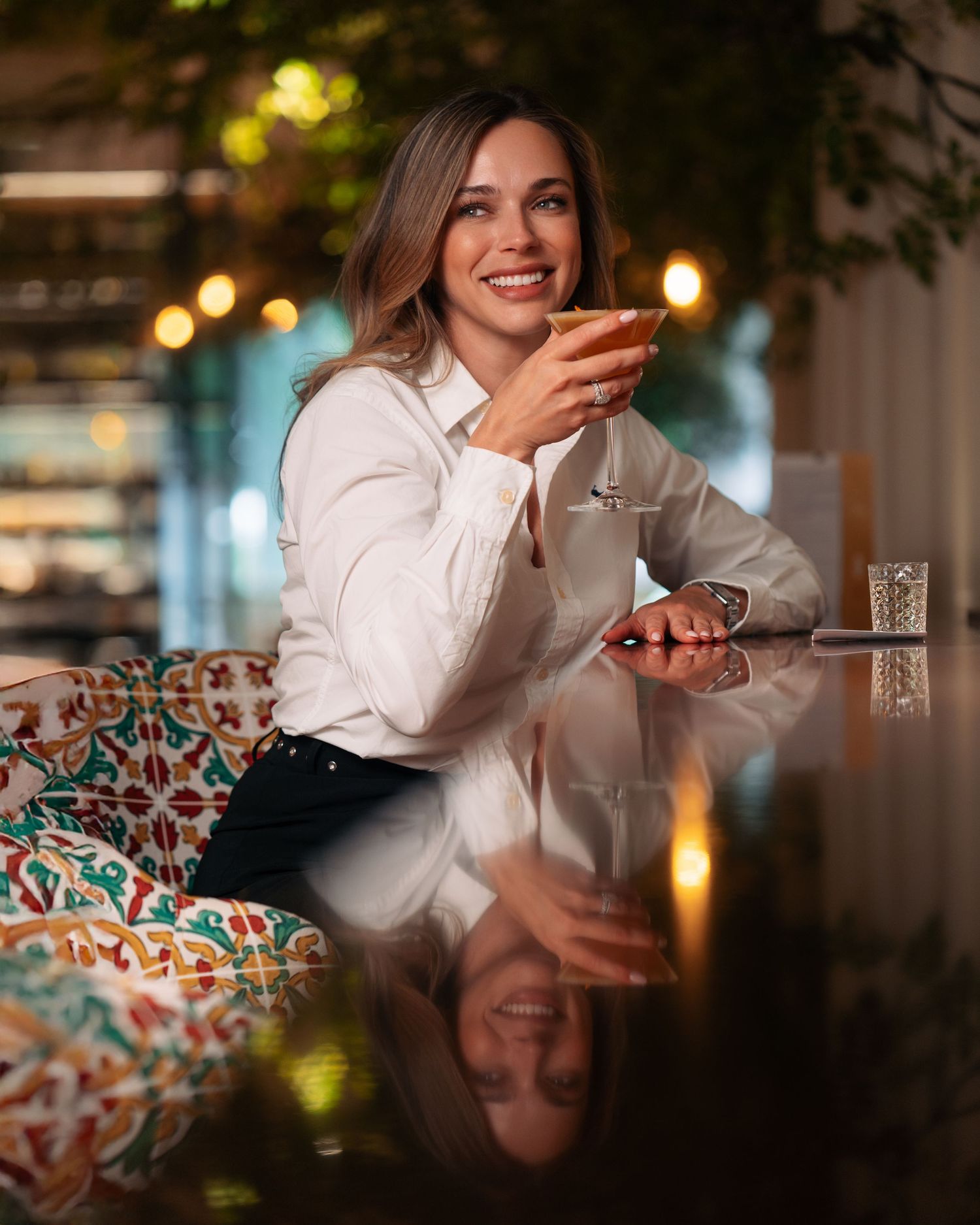 Smiling woman holding a cocktail at a reflective marble table in an elegantly lit restaurant.