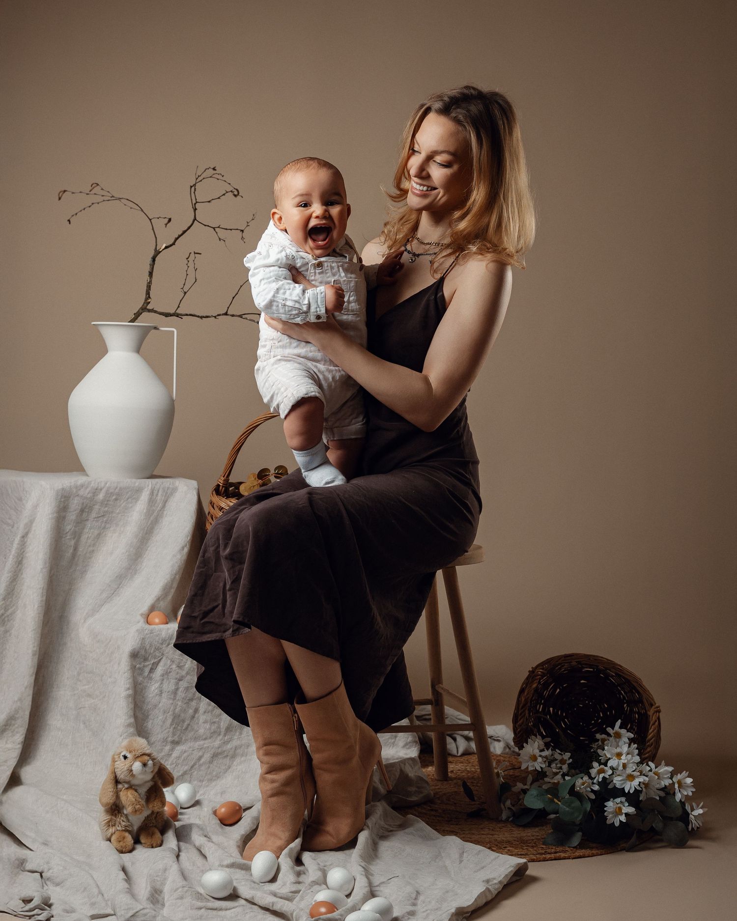 Smiling mother holds laughing baby in a studio portrait with elegant boho decor.