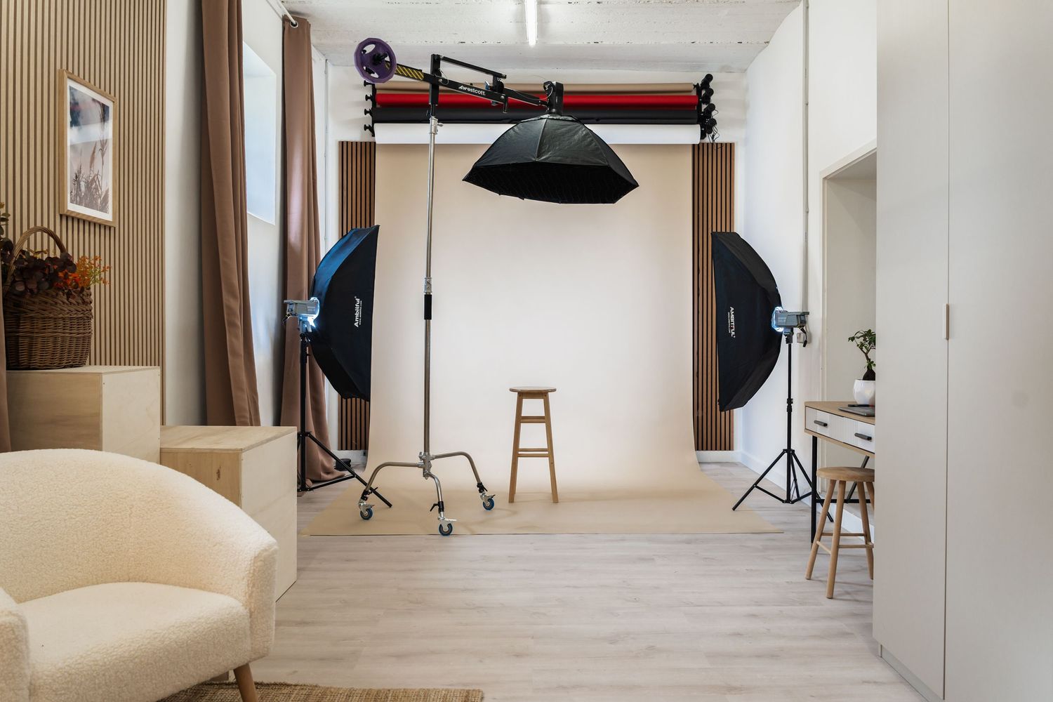 Professional photography studio with white backdrop, softbox lights, stool, and cream armchair on light wood floors.