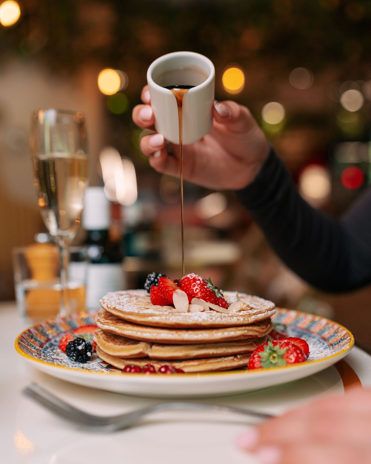 Stack of fluffy pancakes topped with fresh berries and syrup being drizzled from a small cup.
