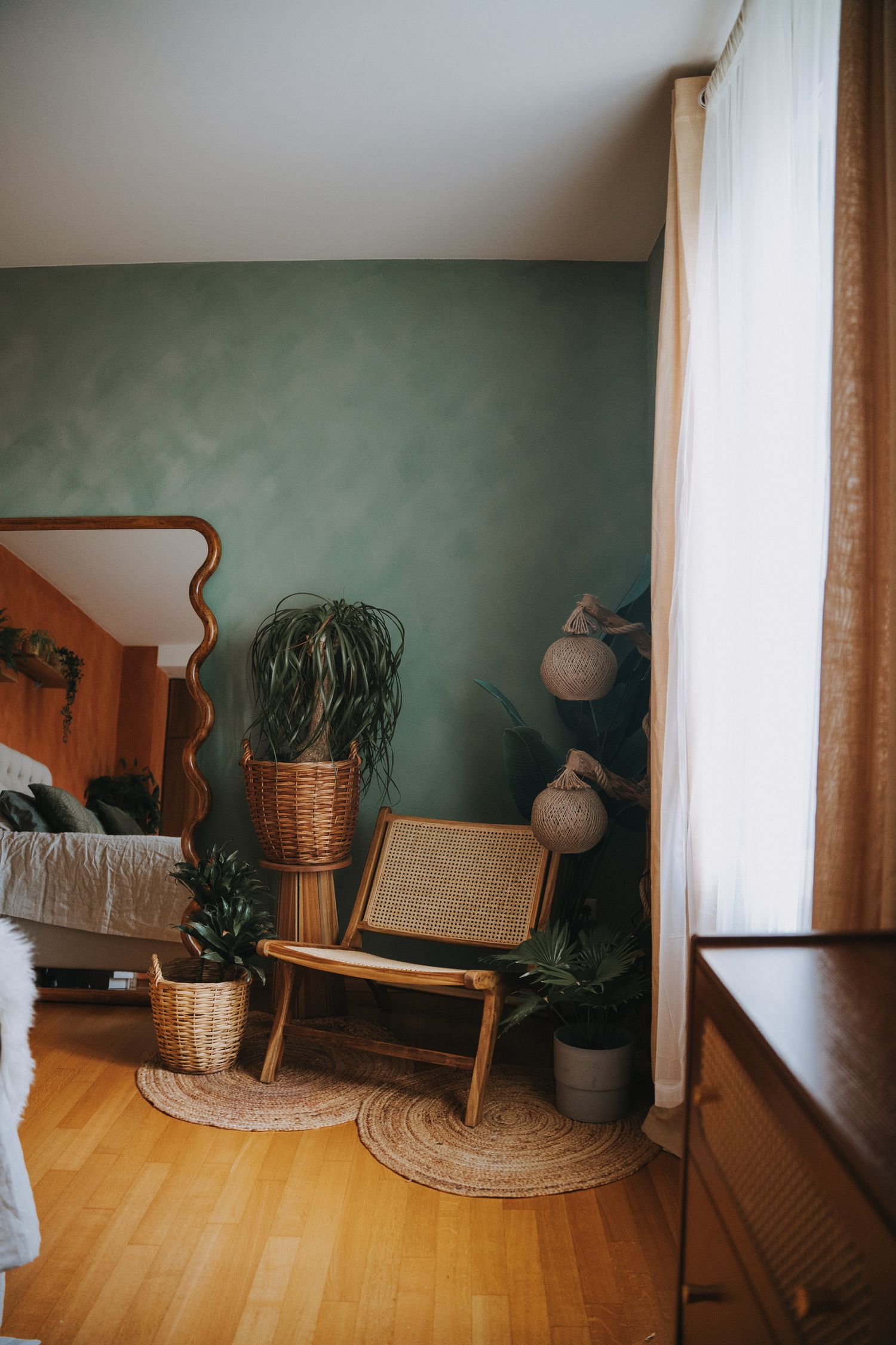 Boho bedroom corner with rattan chair, green plants, sage walls, wood floors, and a wavy mirror reflecting a cozy bed.