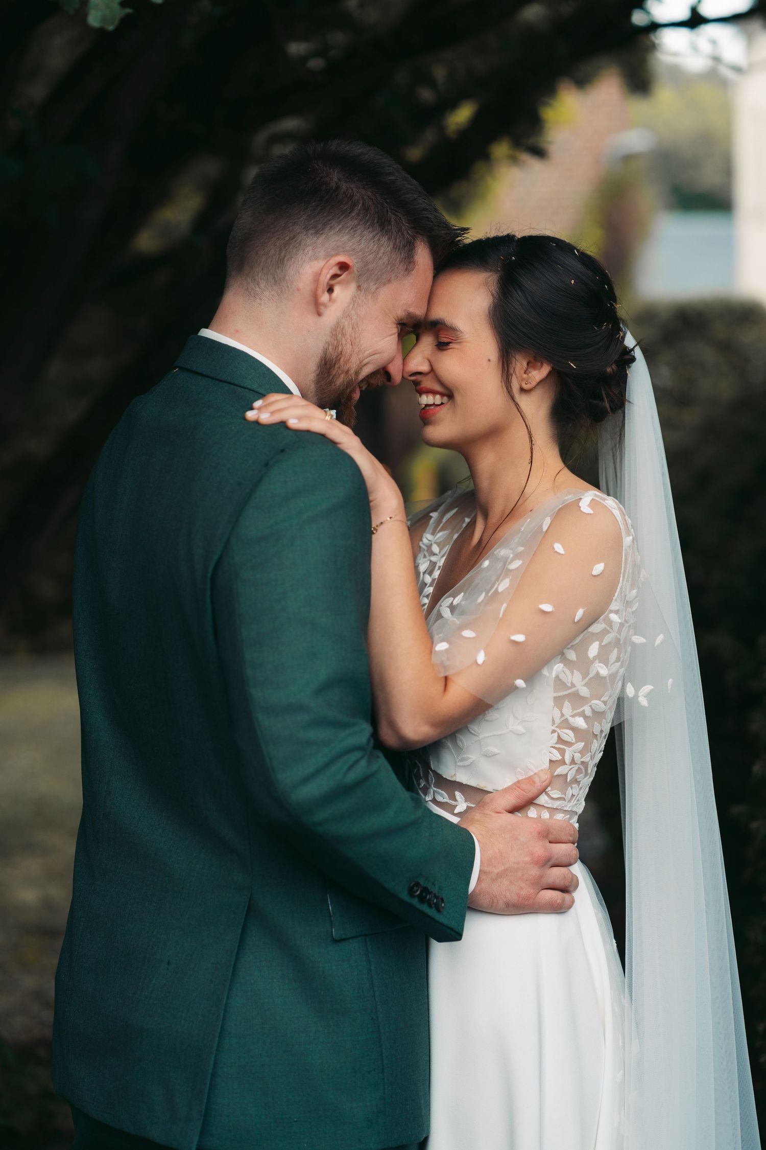 Wedding couple embracing tenderly, bride smiling in embroidered veil dress and groom in dark green suit in garden setting.