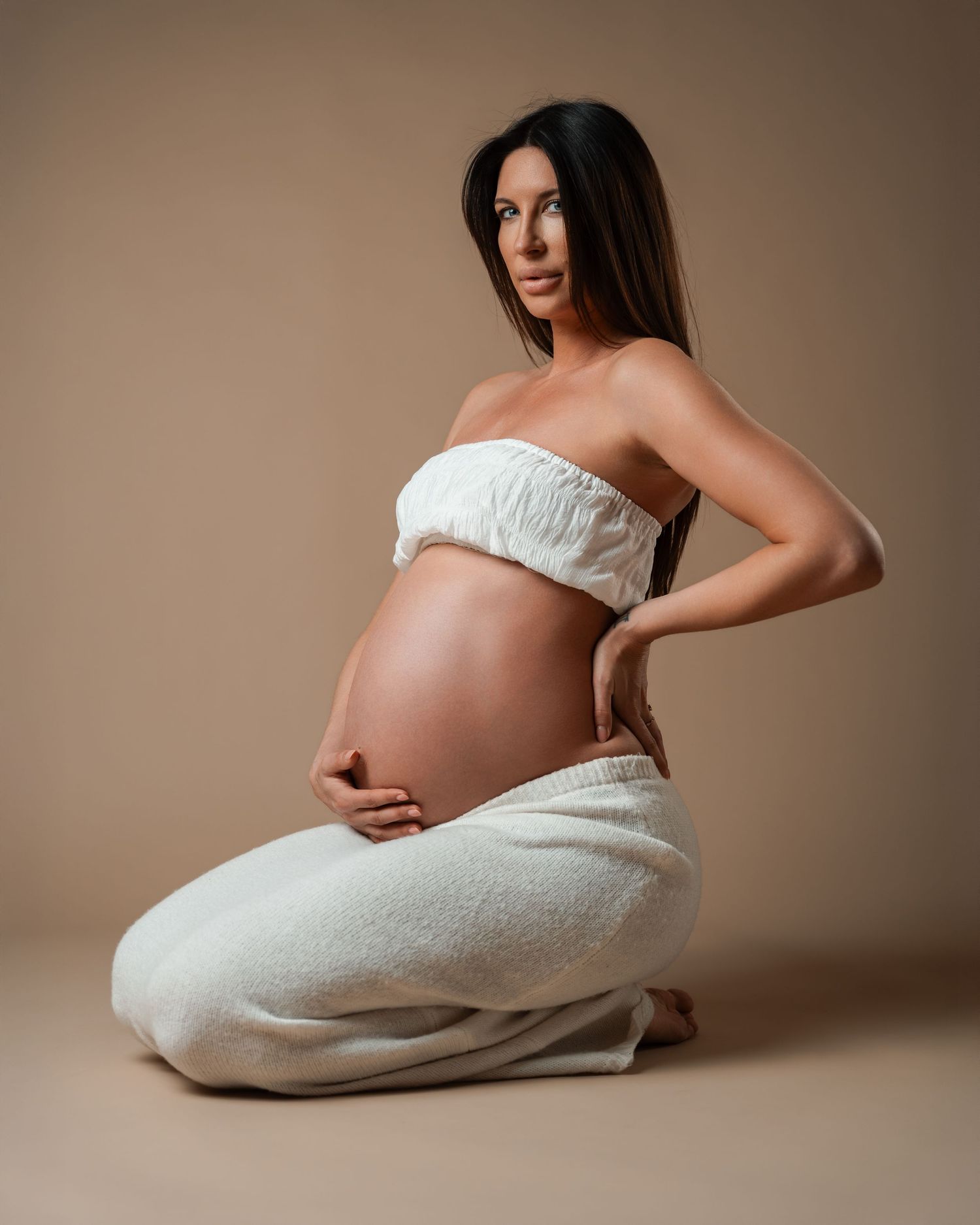 Pregnant woman in white wrap poses gracefully on knees against a neutral beige background.