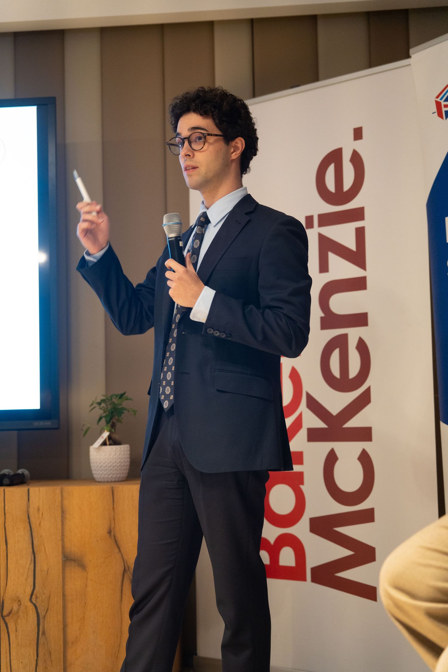 Young male speaker in a suit holding a microphone at a Baker McKenzie corporate event.