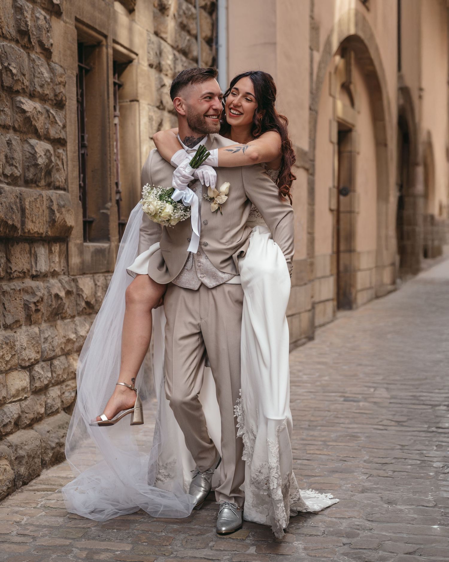 Groom in beige suit lifts smiling bride in white gown holding bouquet on cobblestone street.