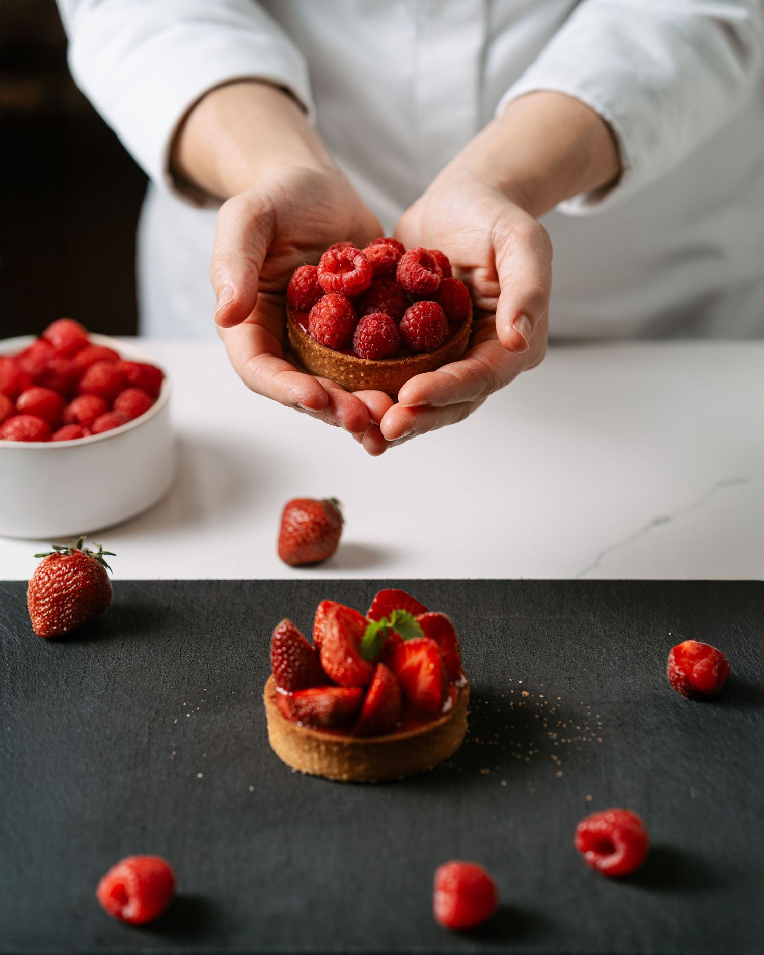 Pastry chef holding a raspberry tart above a strawberry tart on a dark surface.
